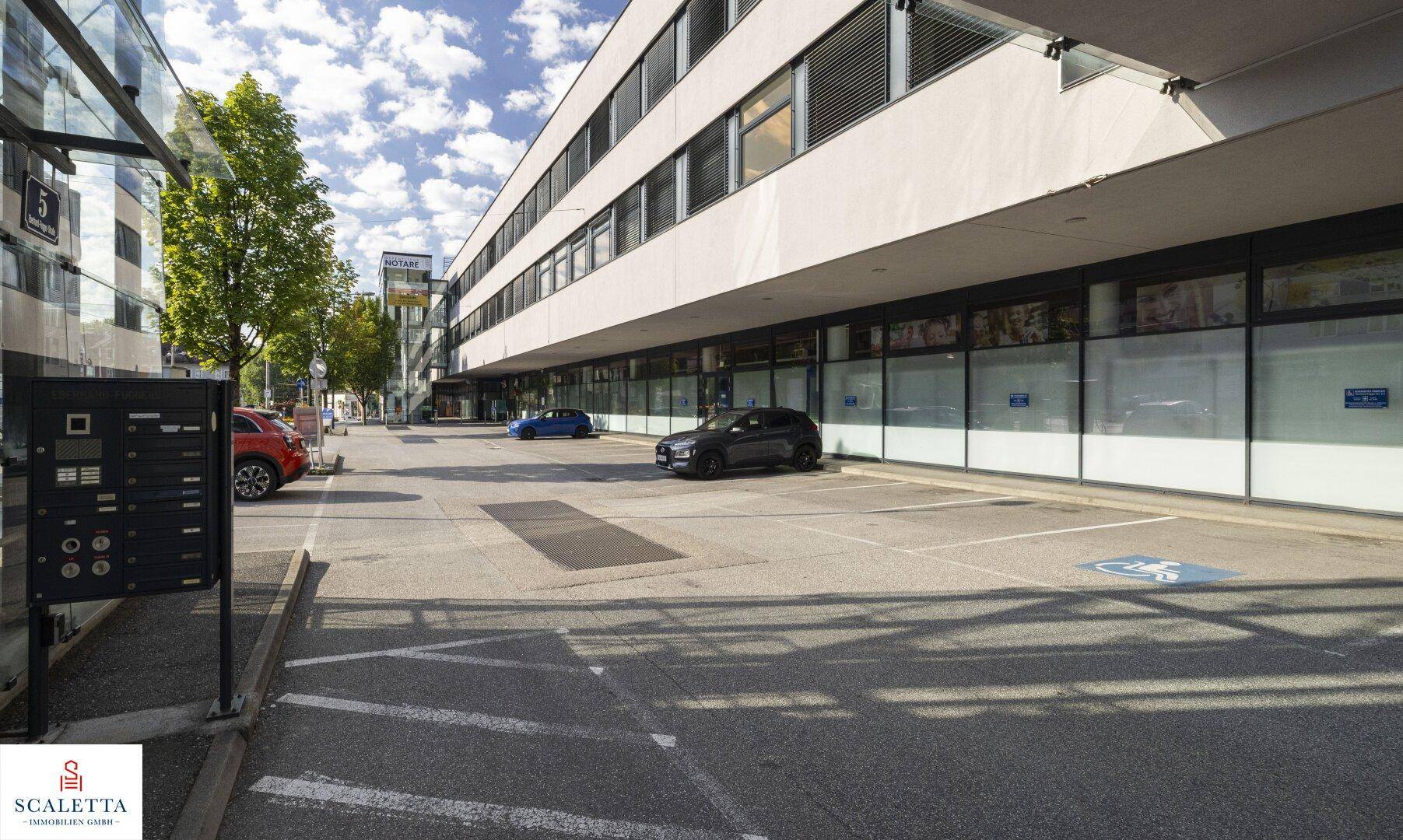 Außenansicht des Geschäftsgebäudes mit Parkplätzen und großen Schaufenstern im Erdgeschoss.
