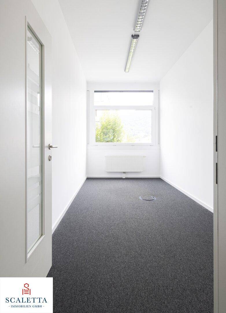 Kleines, helles Büro mit Teppichboden, weißen Wänden und einem Fenster mit Blick ins Grüne.