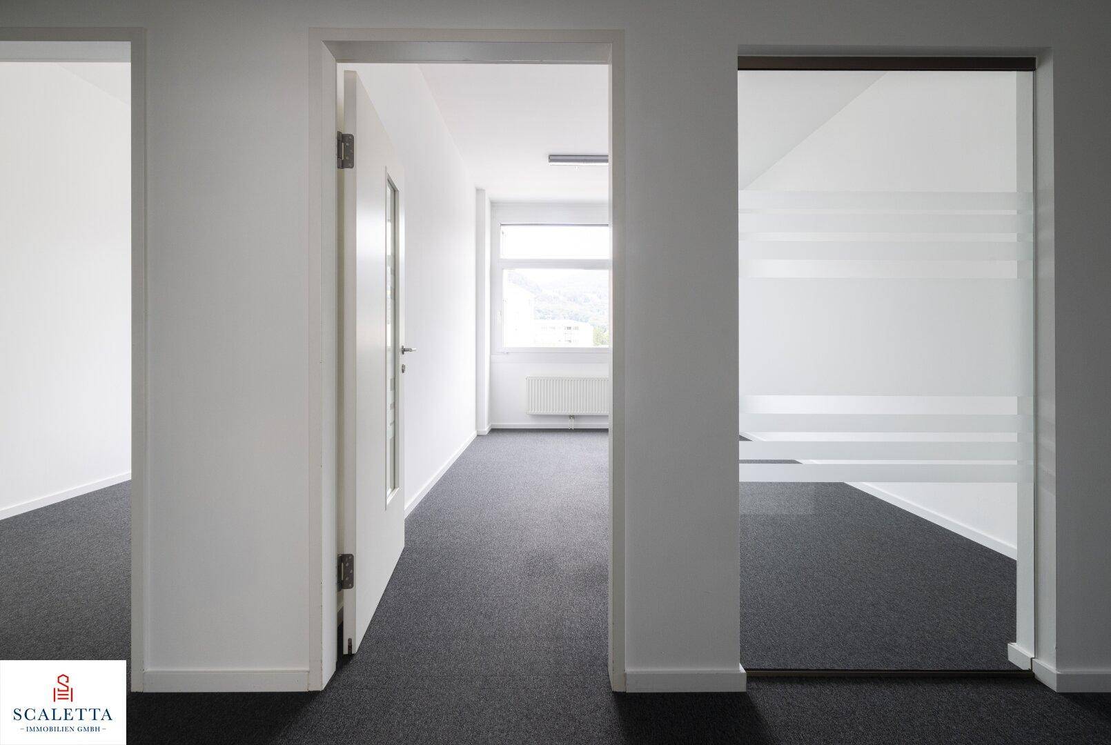 Blick von einem Flur in ein helles Büro mit Teppichboden, weißen Wänden und einem Fenster.