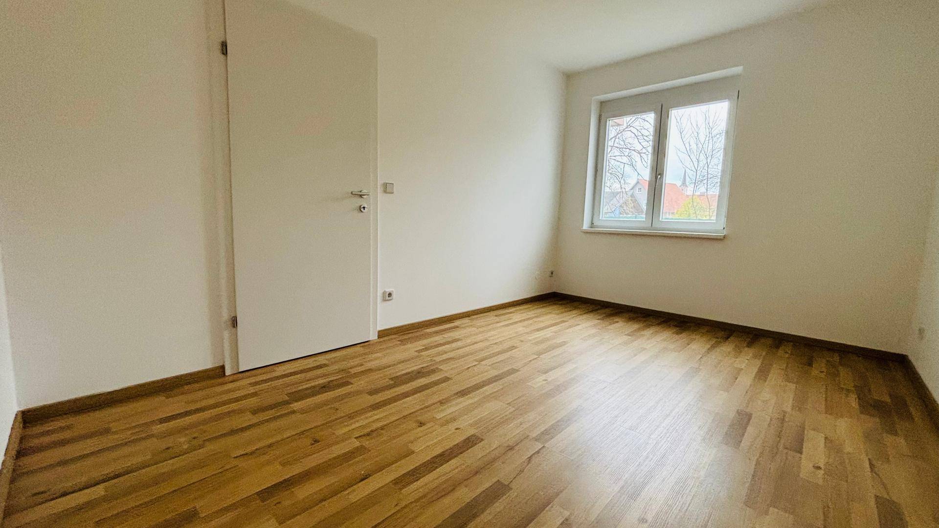 Ruhiges Schlafzimmer mit hellem Parkettboden und großem Fenster.