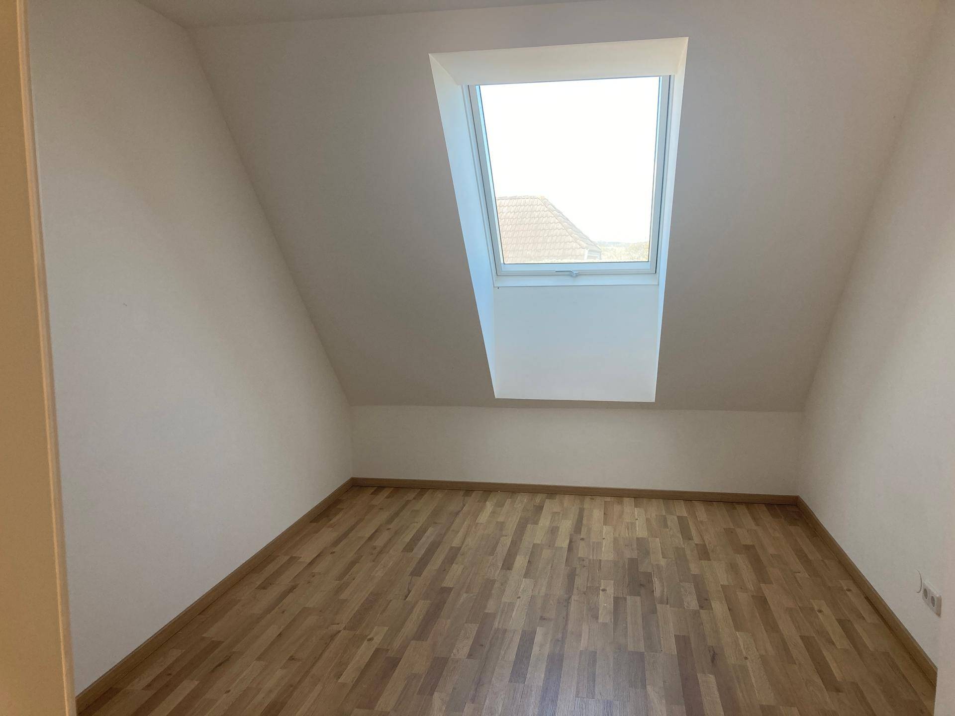 Gemütlicher Dachraum mit einem großen Dachfenster, Holzboden und schrägen Wänden für viel Tageslicht.