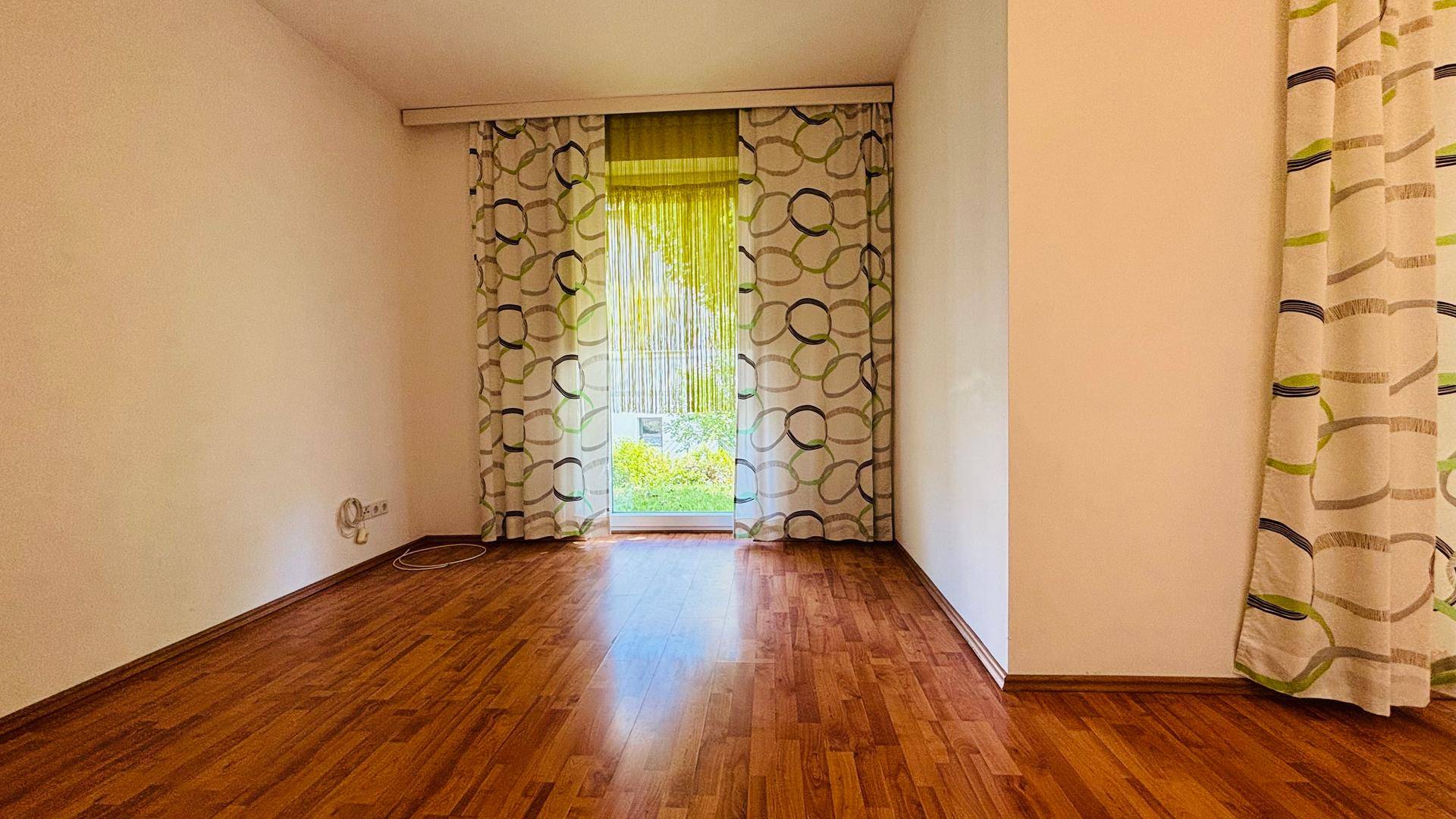 Leeres Zimmer mit Holzboden und großem Fenster, das viel Tageslicht hereinlässt.