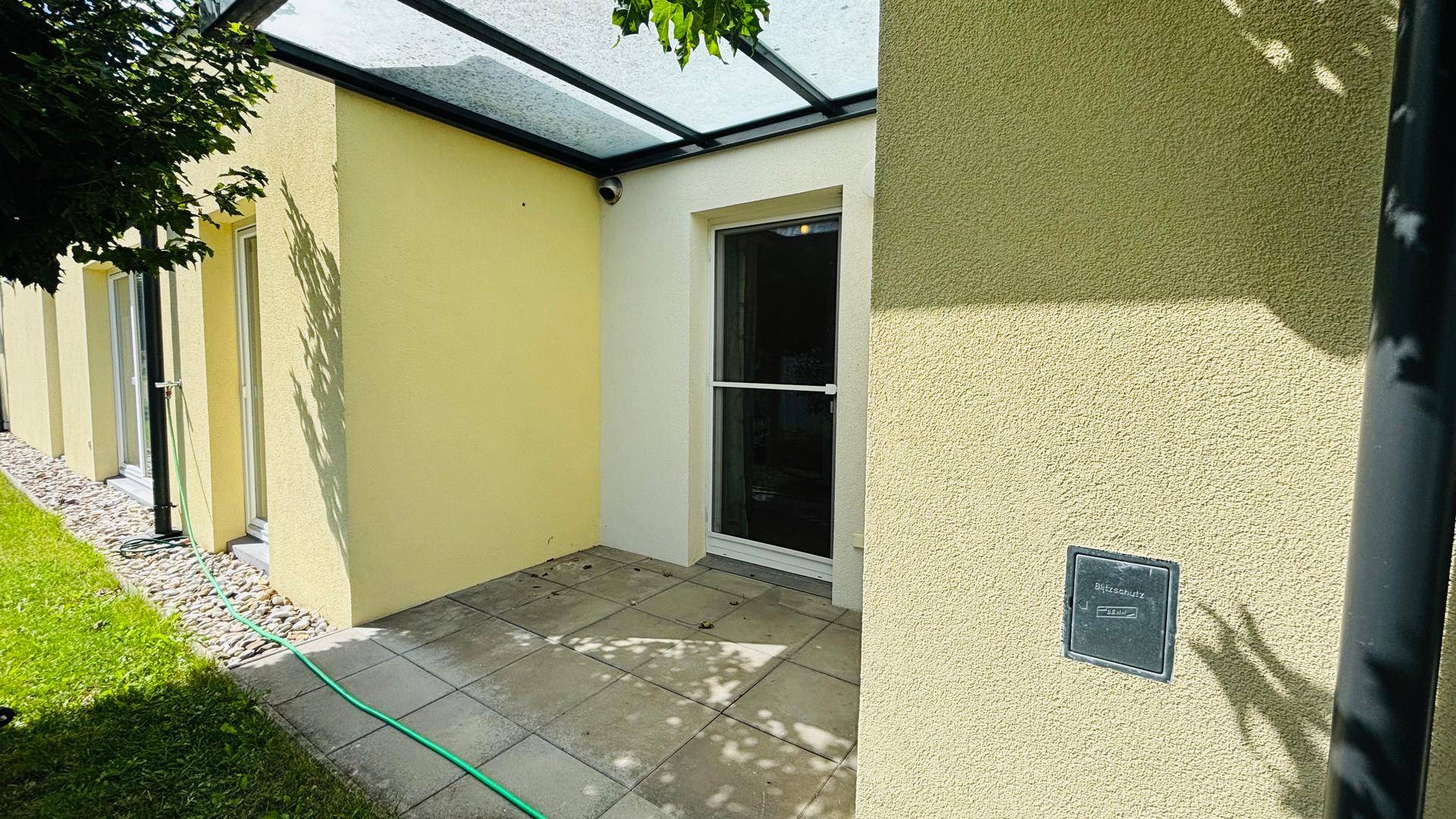 Gepflasterte Terrasse mit Glasüberdachung und Zugang zum grünen Gartenbereich.