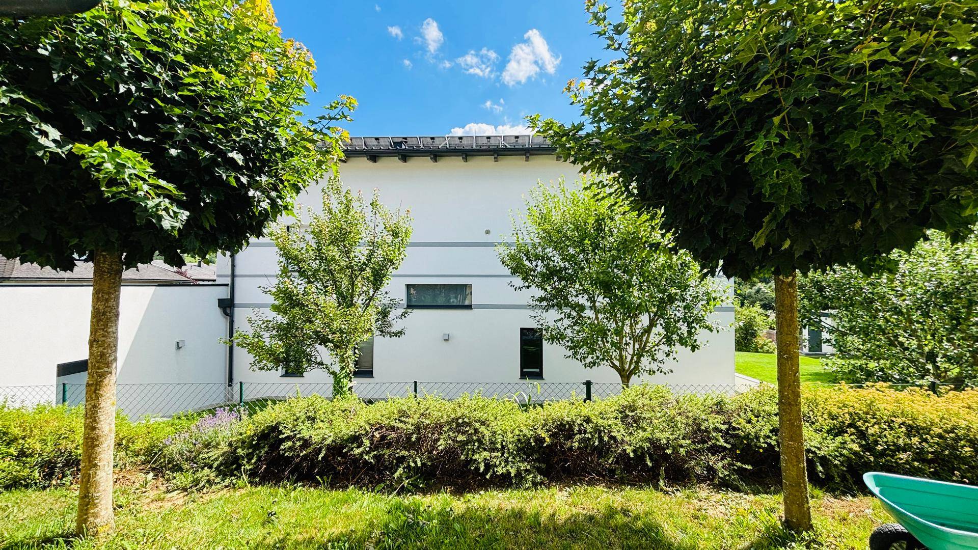 Moderne Gebäudefassade umgeben von gepflegten Bäumen und Sträuchern unter blauem Himmel.