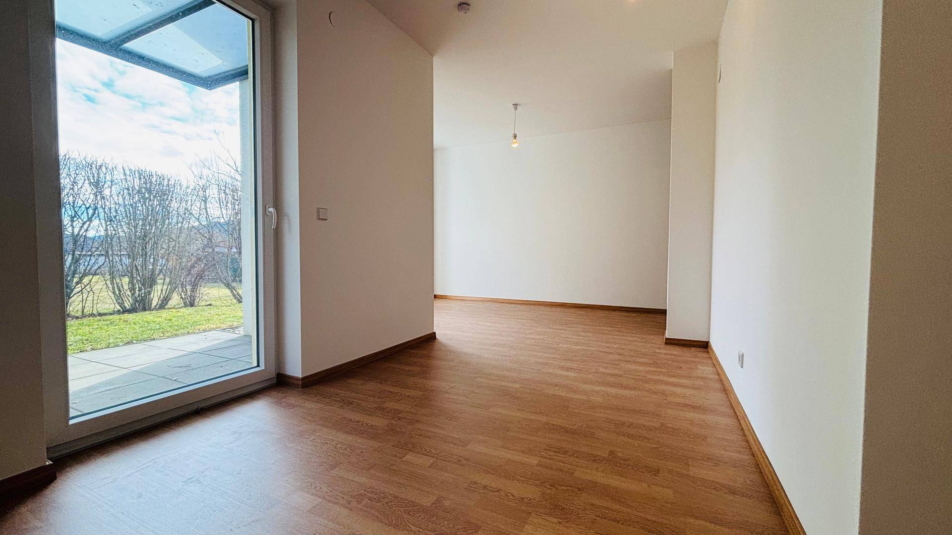 Offener Wohnbereich mit Holzboden und großer Terrassentür, die den Blick ins Grüne freigibt.