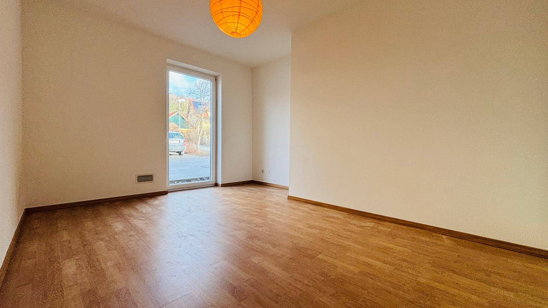 Helles Zimmer mit Holzboden und einem Fenster, das den Blick auf die Straße ermöglicht.