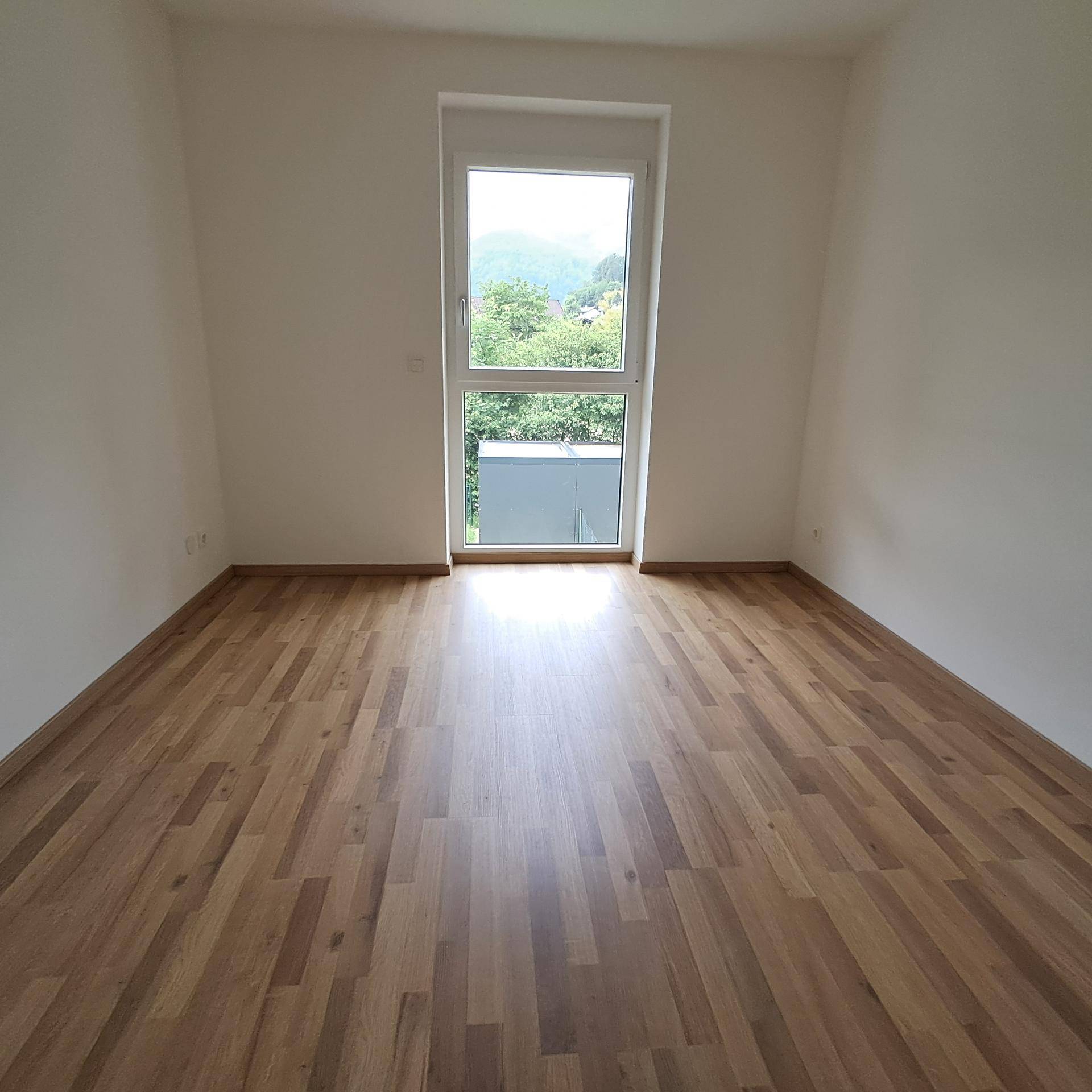 Leeres Schlafzimmer mit Laminatboden und großem Fenster mit Blick ins Grüne.