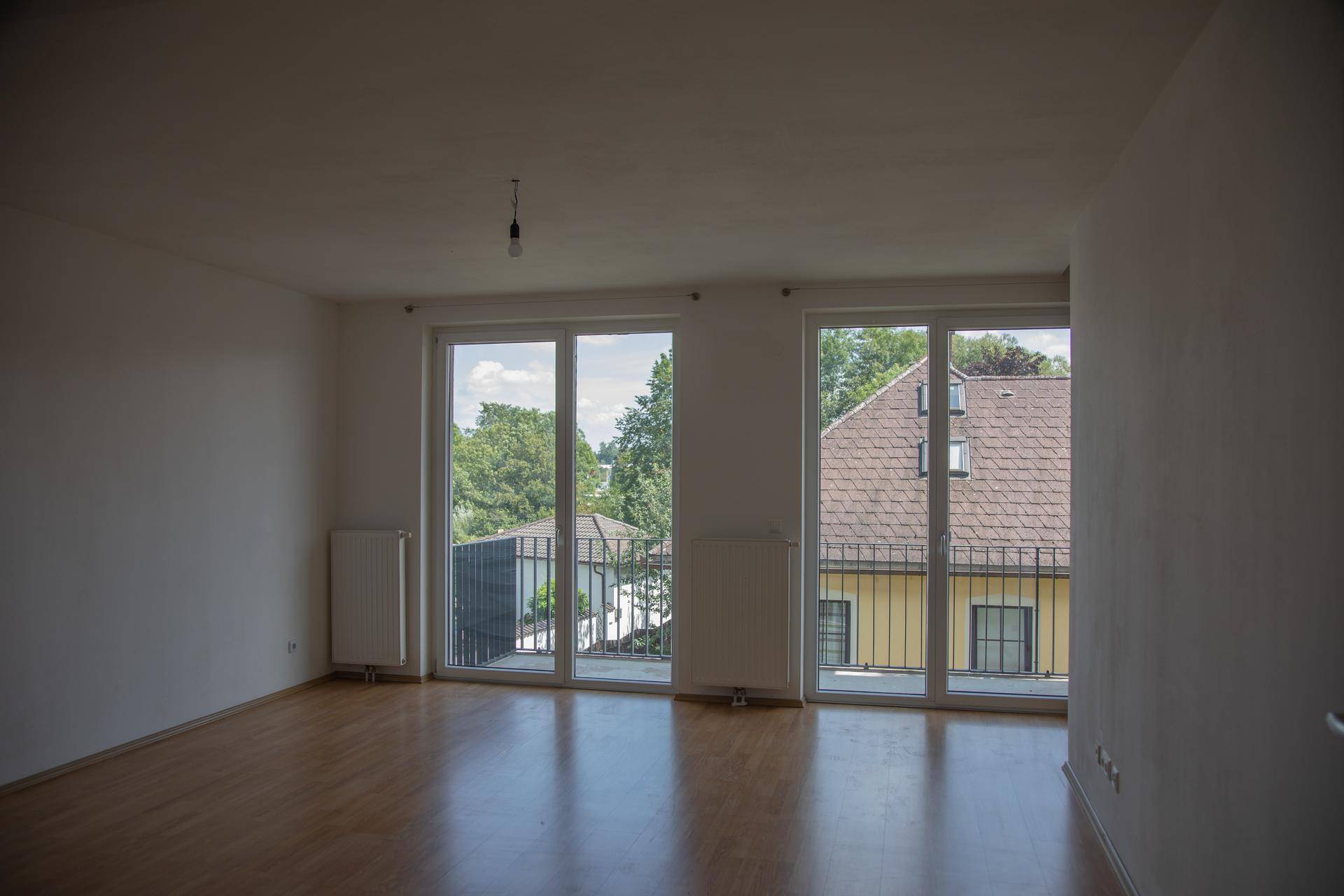 Helles, leeres Wohnzimmer mit Parkettboden und Zugang zu einem Balkon mit Blick ins Grüne.