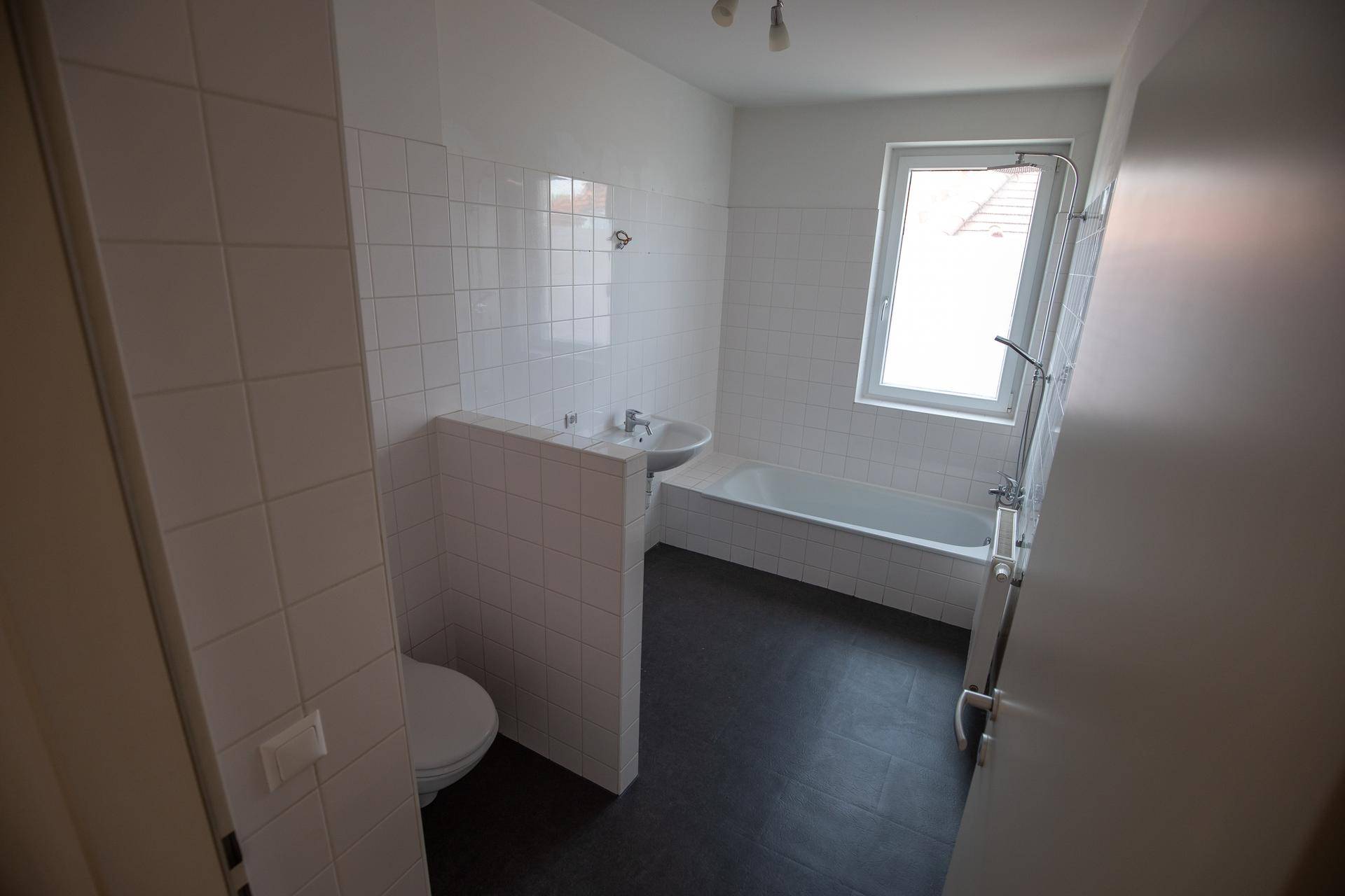 Badezimmer mit weißen Fliesen, Badewanne, Toilette und einem Fenster für Tageslicht.