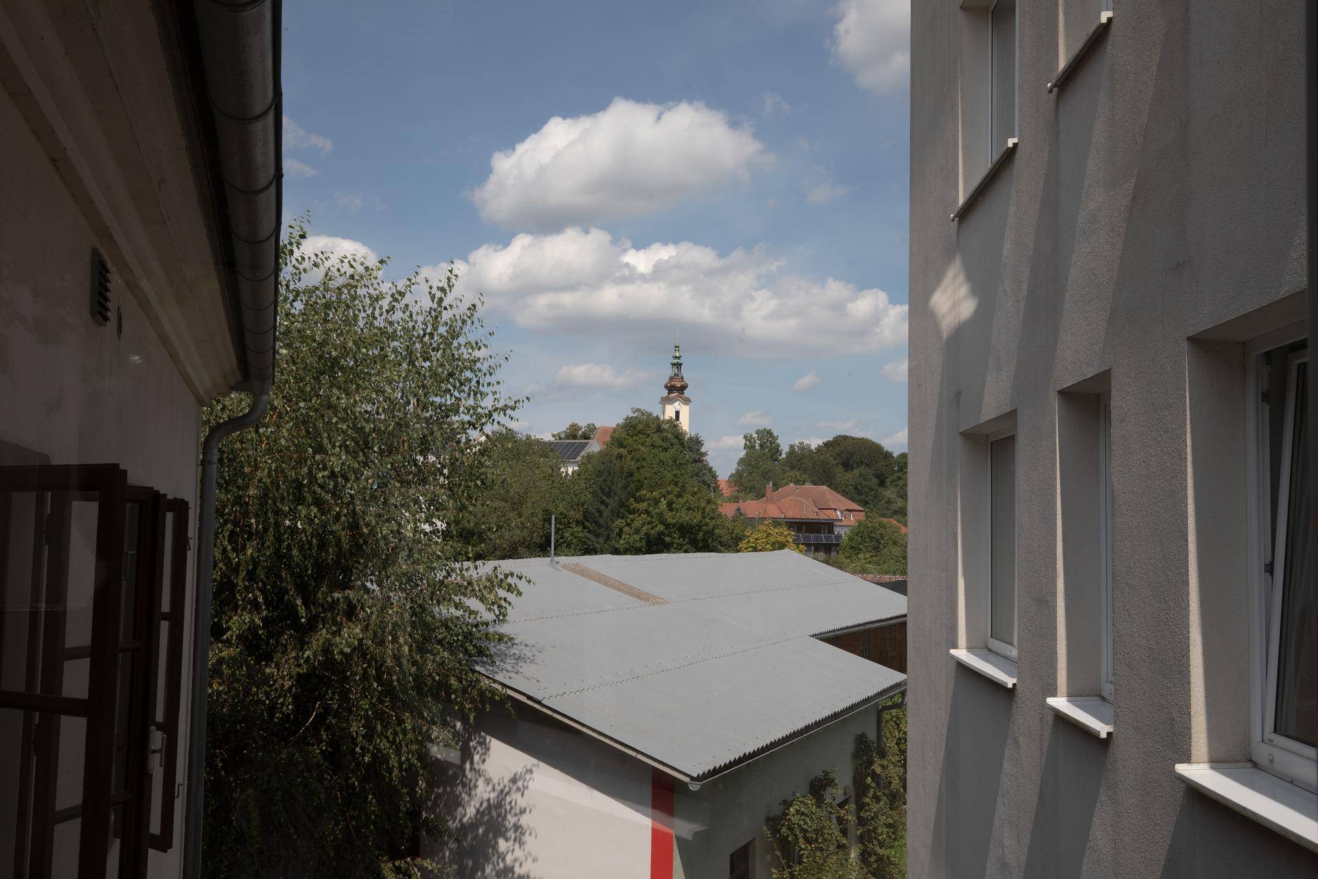 Blick aus dem Fenster auf einen Kirchturm und grüne Bäume unter blauem Himmel.