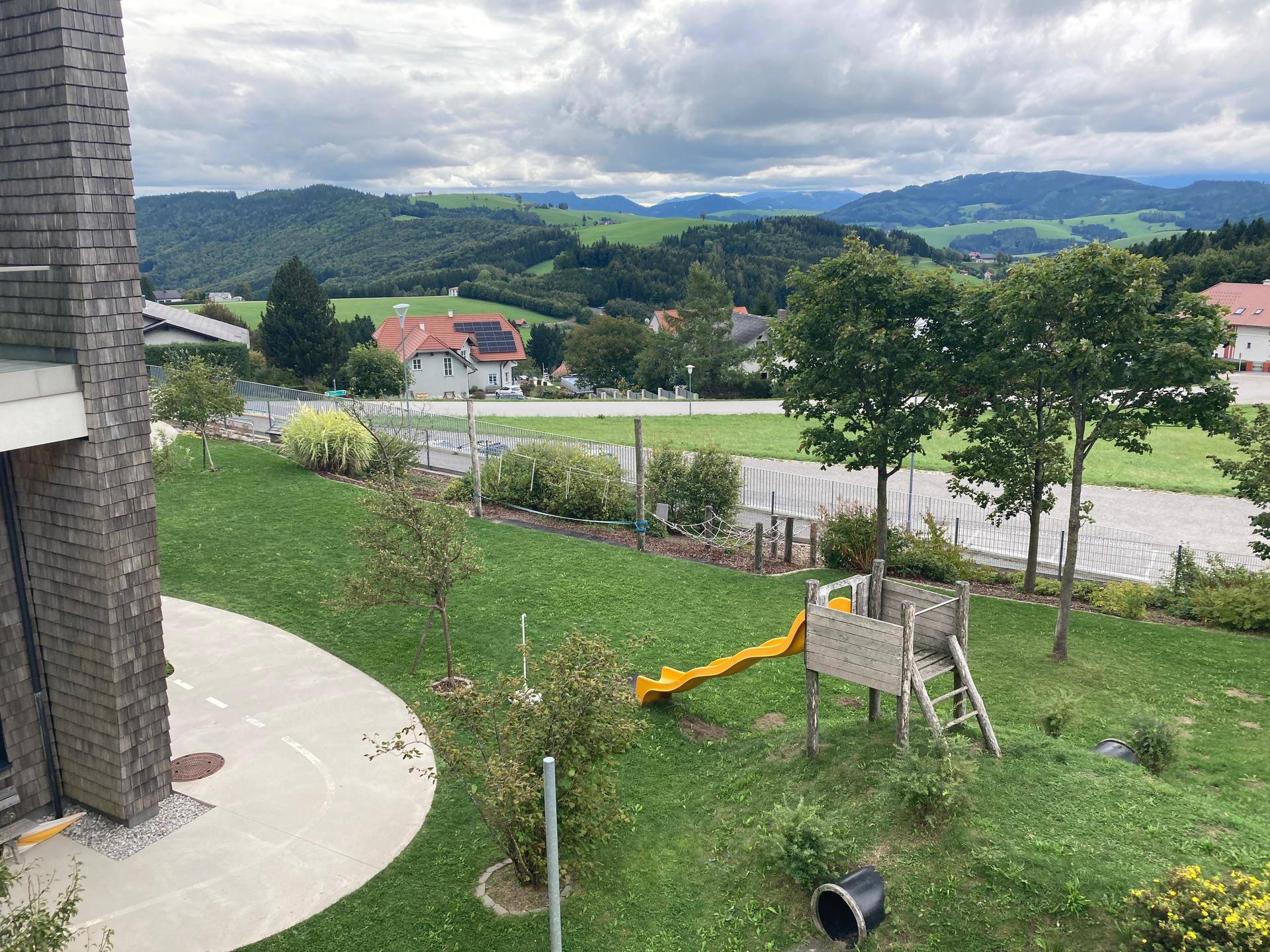Kinderspielplatz mit Rutsche und Schaukel vor einer malerischen Hügellandschaft mit Häusern.