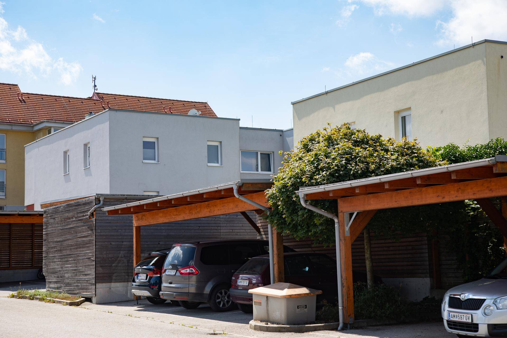 Mehrere Carports aus Holz bieten überdachte Parkplätze vor modernen Wohngebäuden.