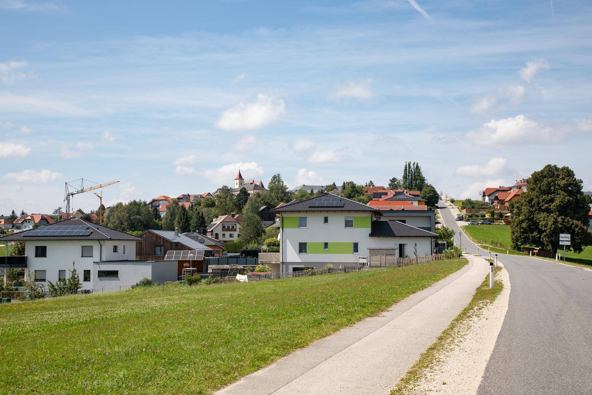 Landstraße führt durch eine ländliche Siedlung mit modernen Häusern und einem Kirchturm in der Ferne.