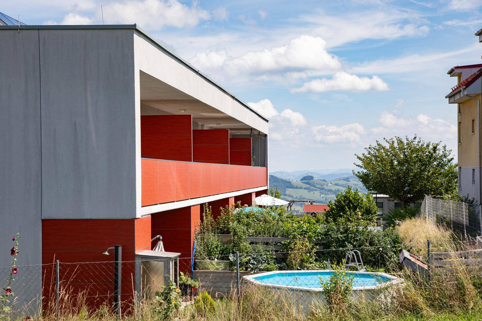Moderne Wohnanlage mit roter Fassade, Gartenbereich und einem Aufstellpool mit Bergblick.