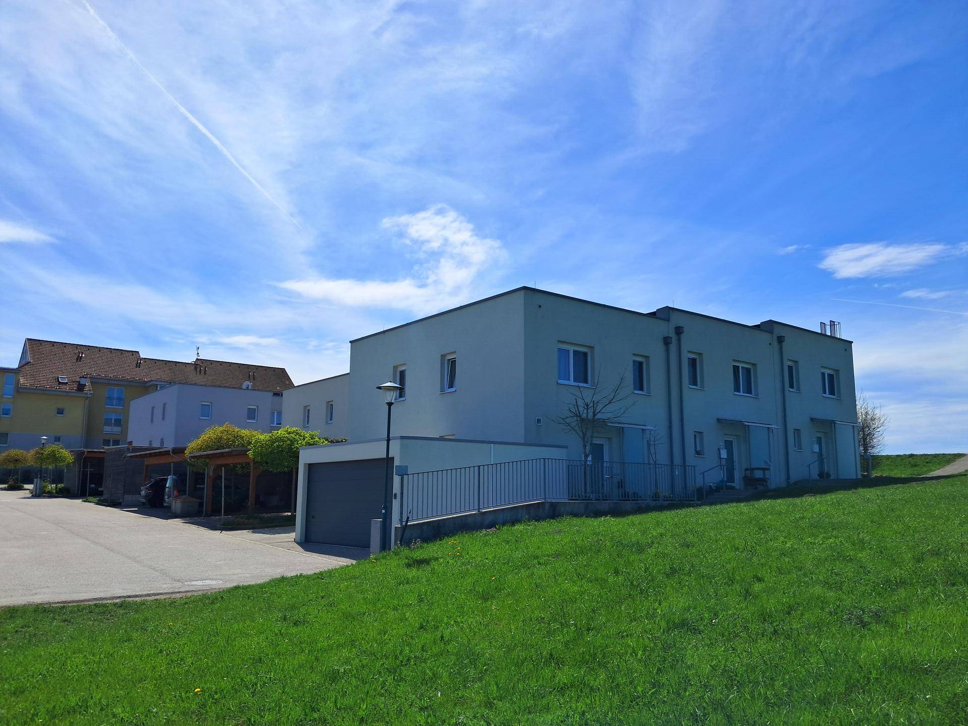 Moderne Wohngebäude mit Garagen und Carports, umgeben von grünen Flächen unter blauem Himmel.