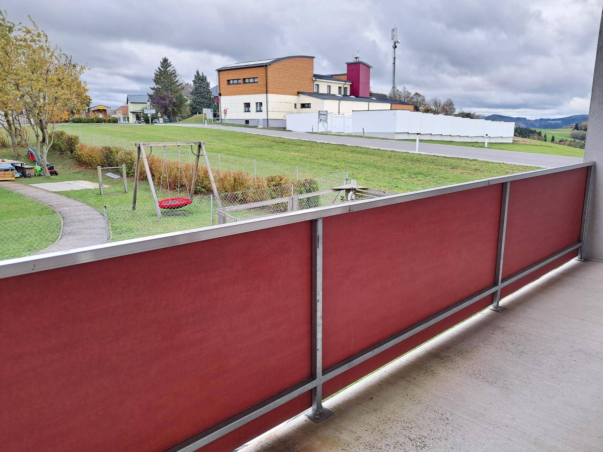 Balkon mit roter Brüstung und Blick auf einen Spielplatz und die umliegende Landschaft.