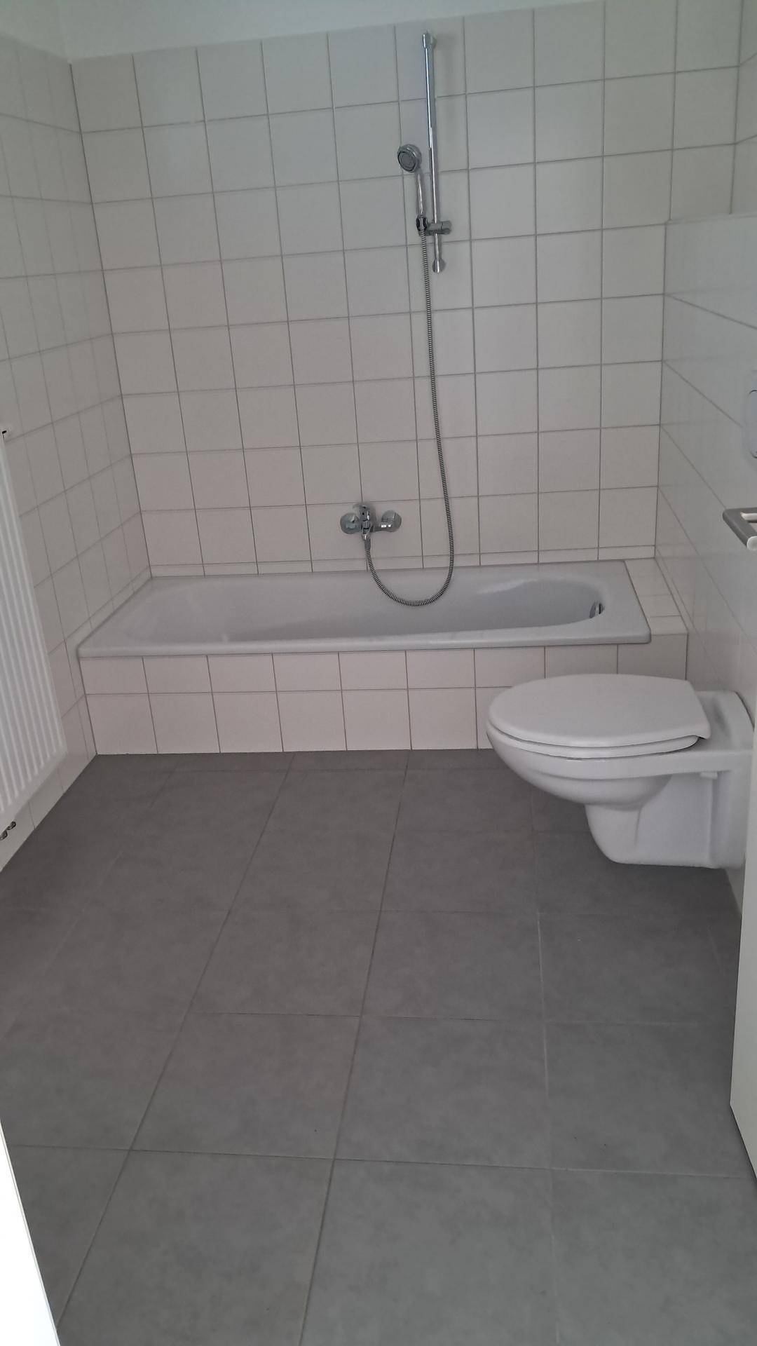 Badezimmer mit Badewanne, Wand-WC und grauen Bodenfliesen in neuwertigem Zustand.