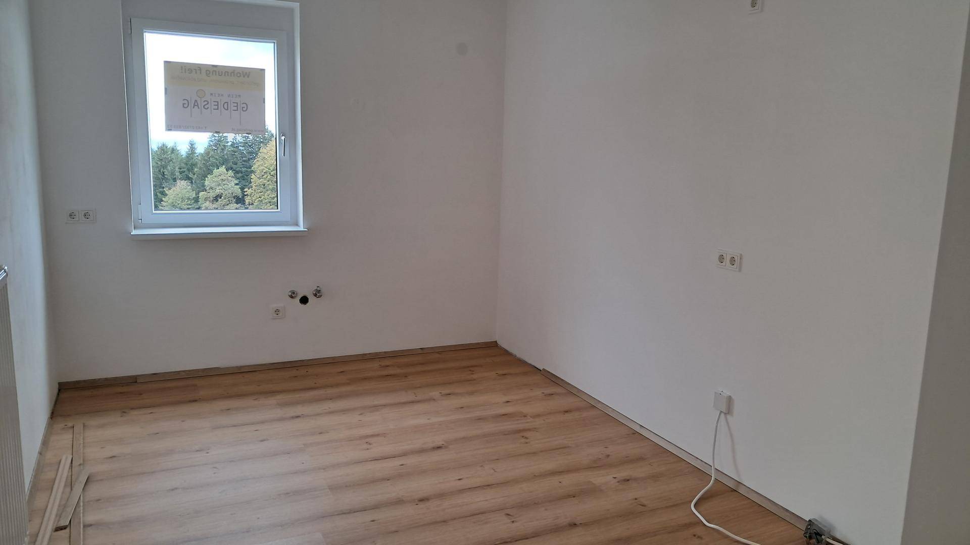 Leeres Schlafzimmer mit Laminatboden, Heizkörper und Fenster mit Blick ins Grüne.