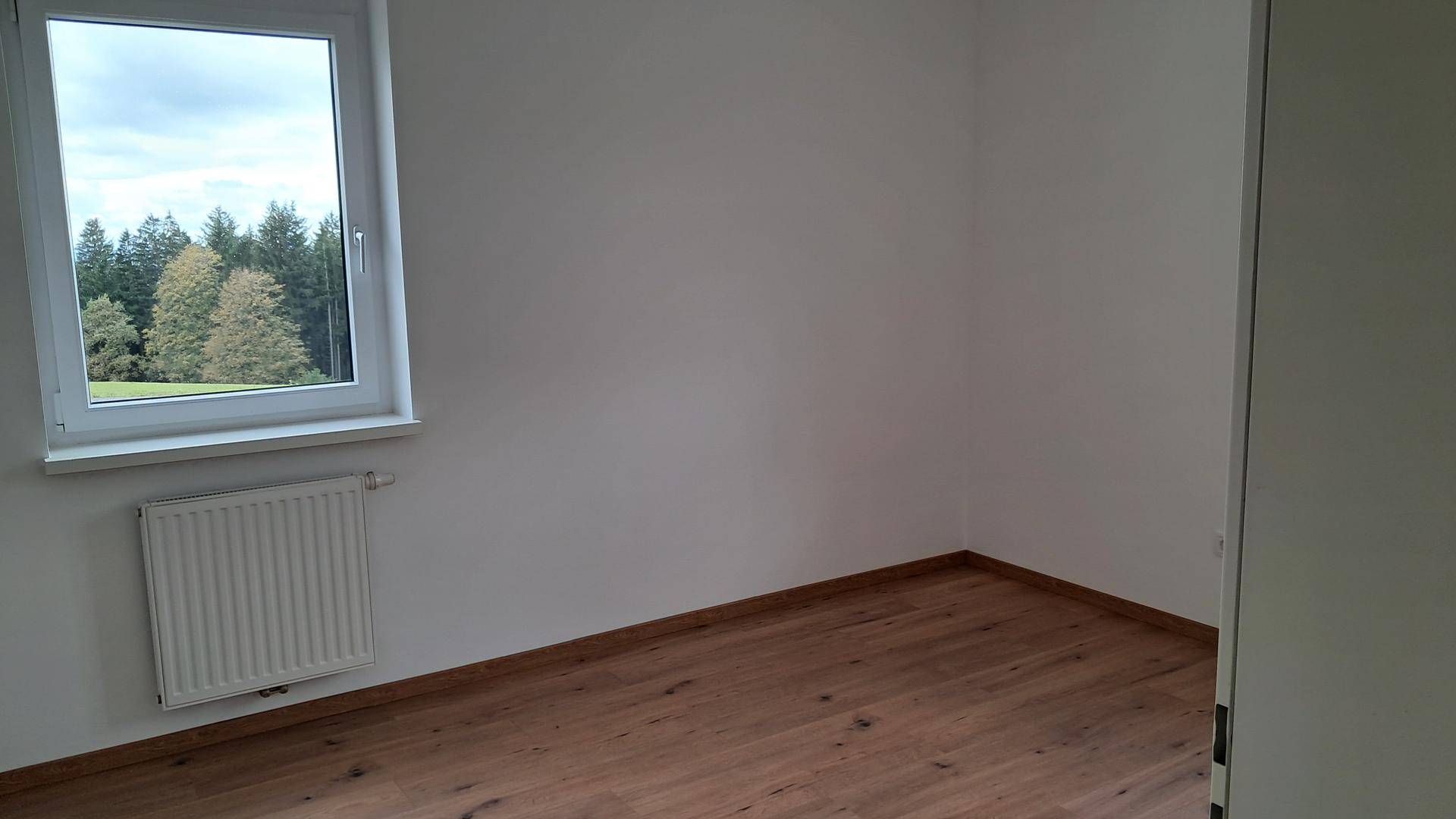 Leeres Schlafzimmer mit Laminatboden, Heizkörper und Fenster mit Blick auf Bäume.
