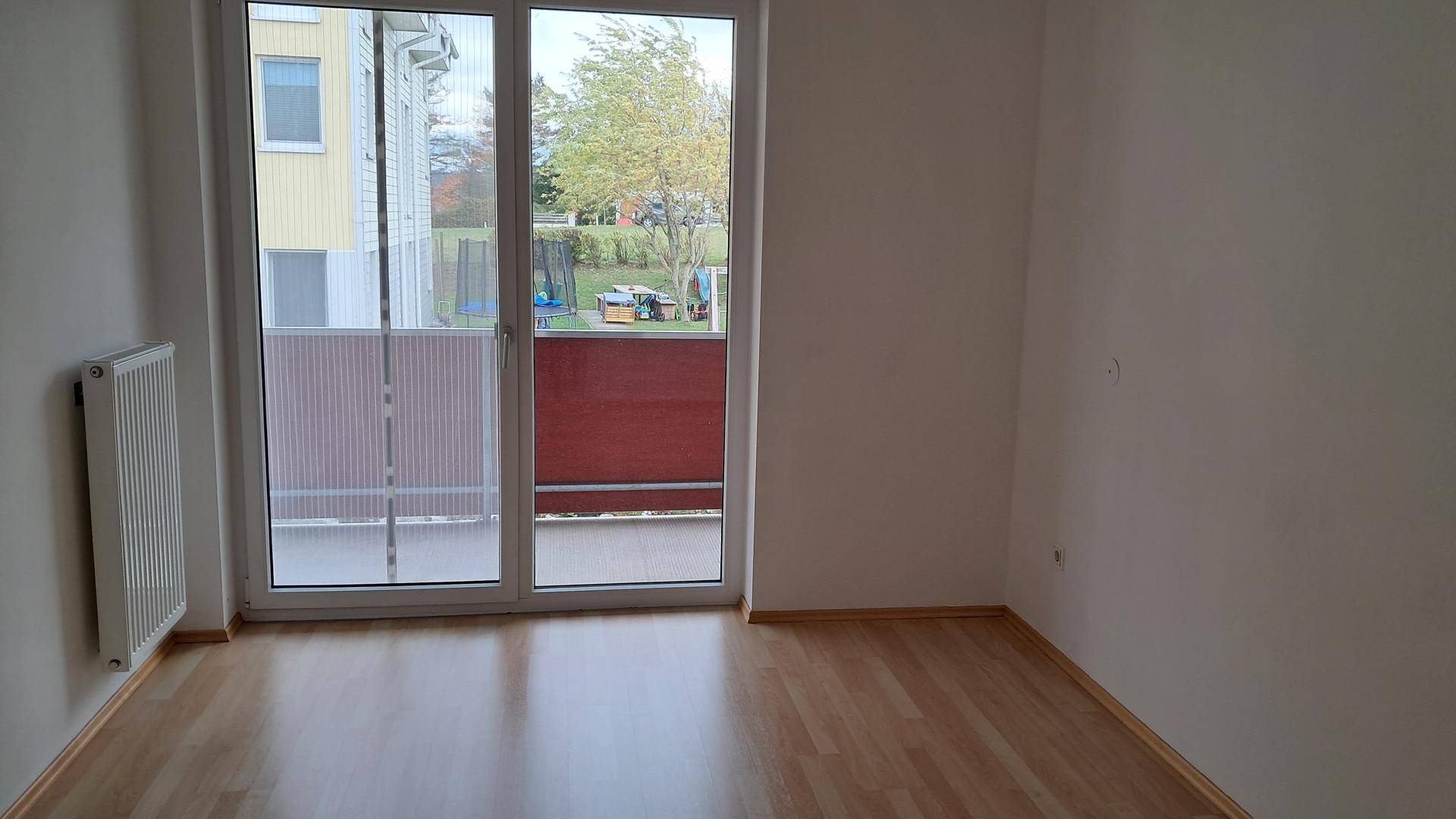 Leerer Raum mit Laminatboden, Heizkörper und Zugang zu einem Balkon mit roter Brüstung.