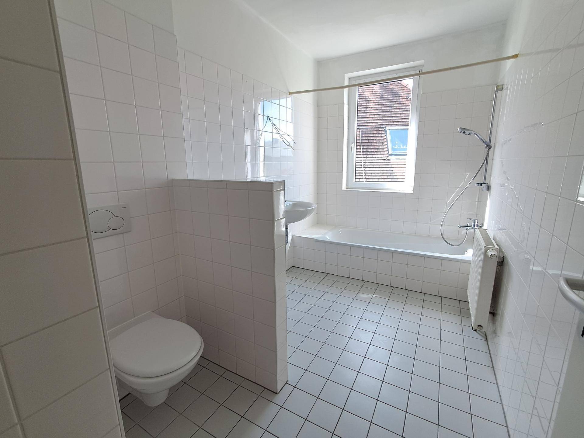 Helles Badezimmer mit weißen Fliesen, Badewanne, Toilette und einem Fenster für Tageslicht.