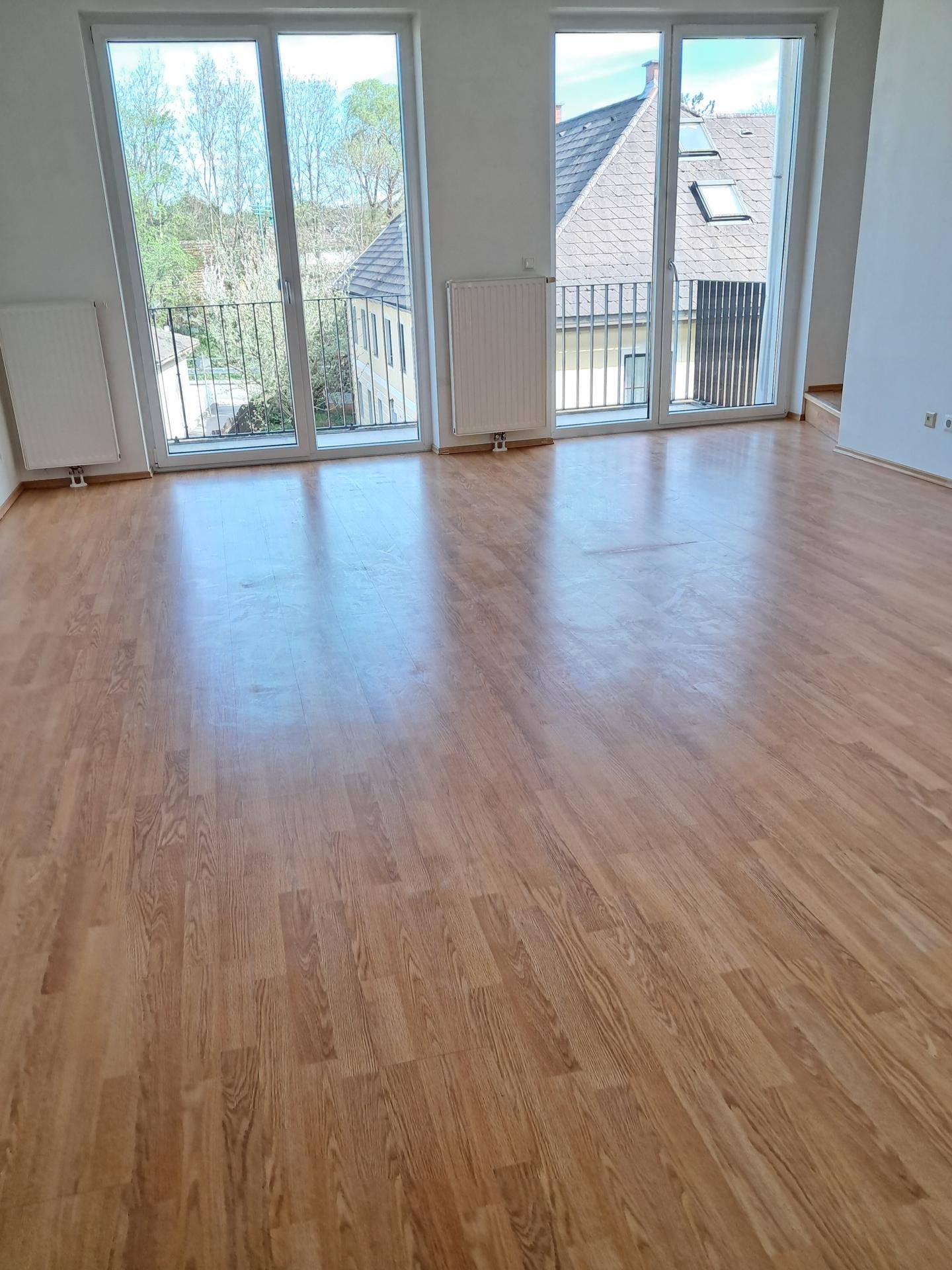 Heller Wohnbereich mit Laminatboden und großen Fenstern, die Zugang zu einem Balkon bieten.