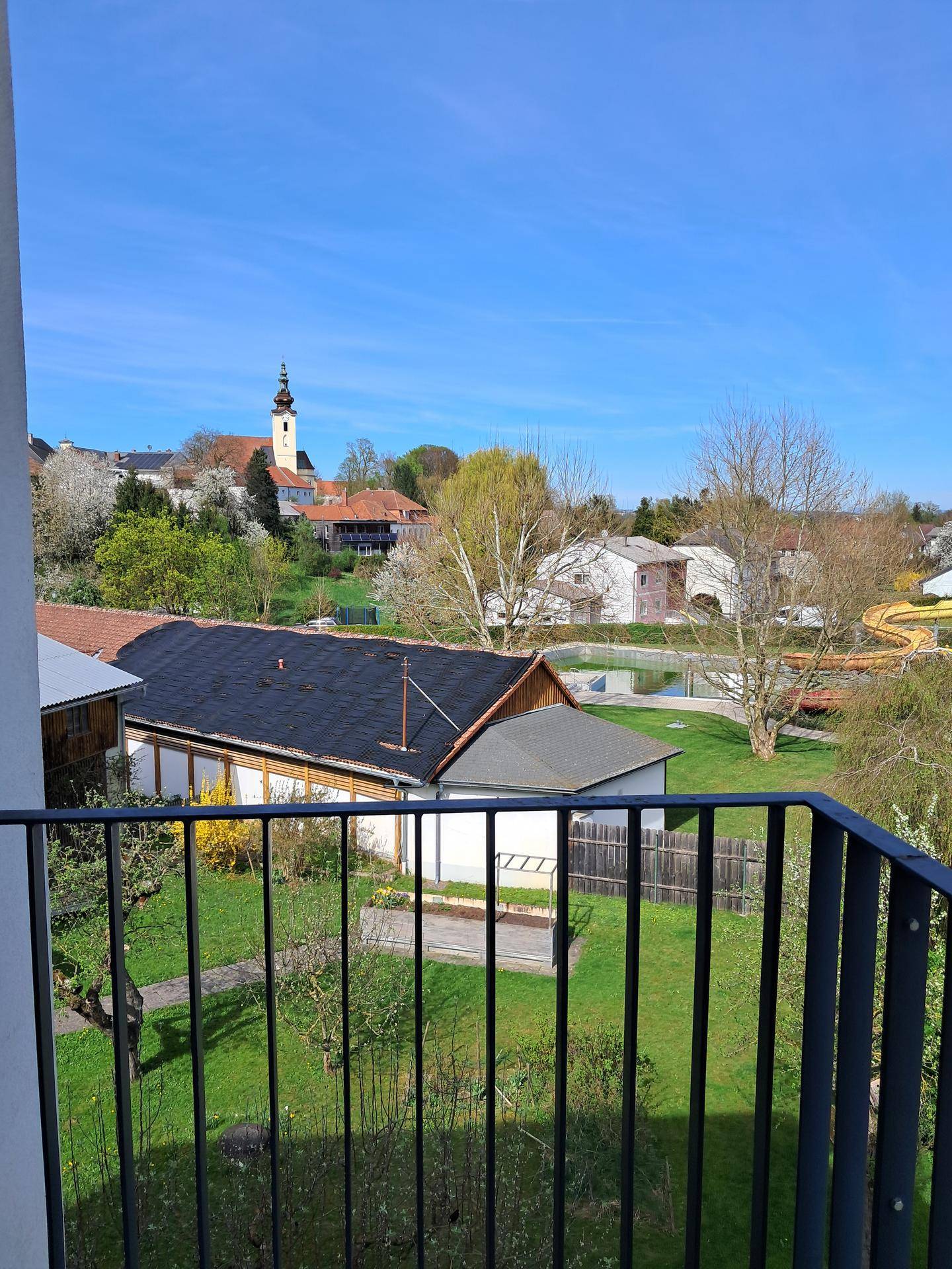 Balkon mit Blick auf eine grüne Umgebung und einen Kirchturm in der Ferne unter blauem Himmel.