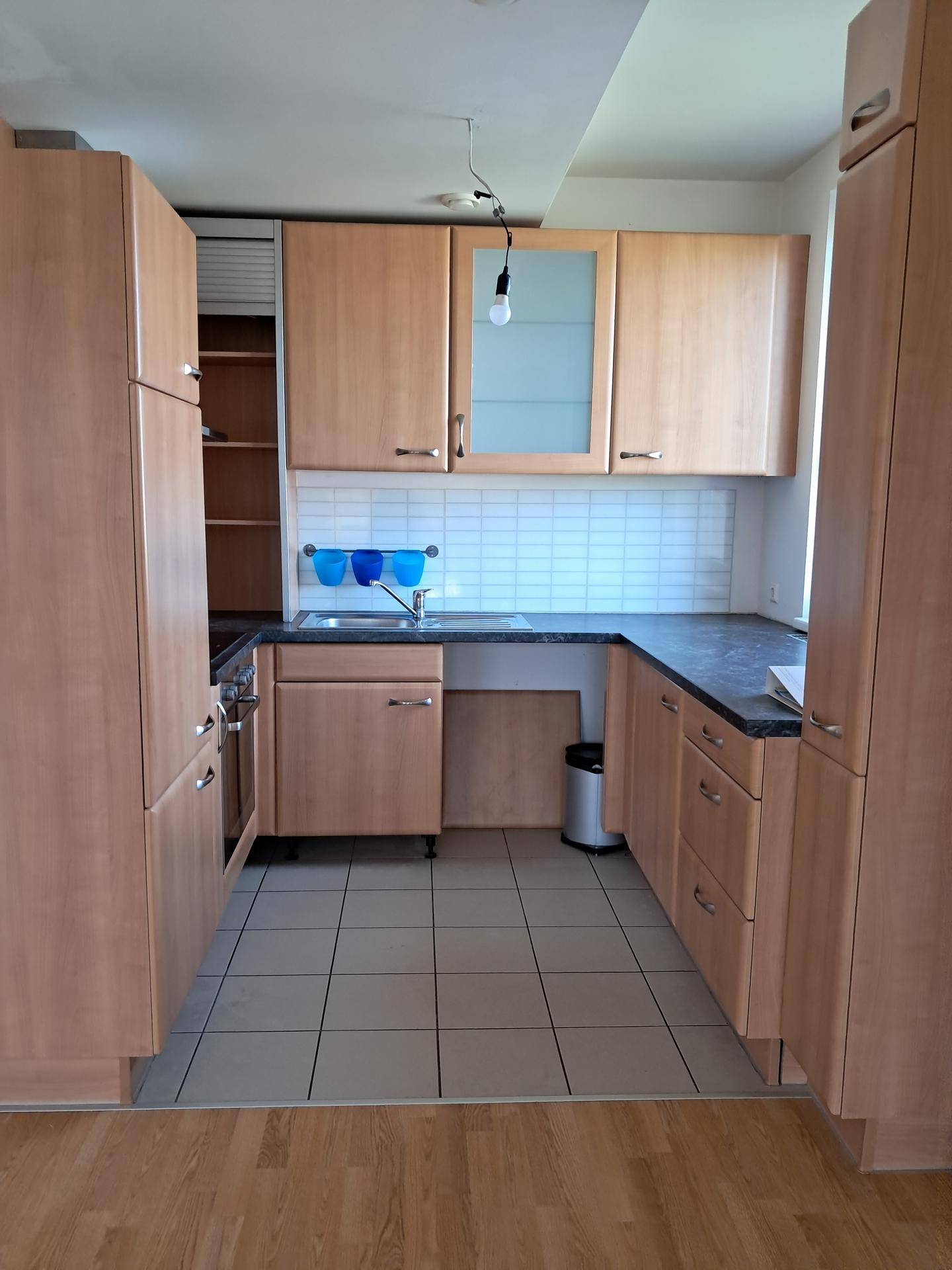 Funktionale Einbauküche mit hellen Holzschränken, dunkler Arbeitsplatte und Fliesenboden.