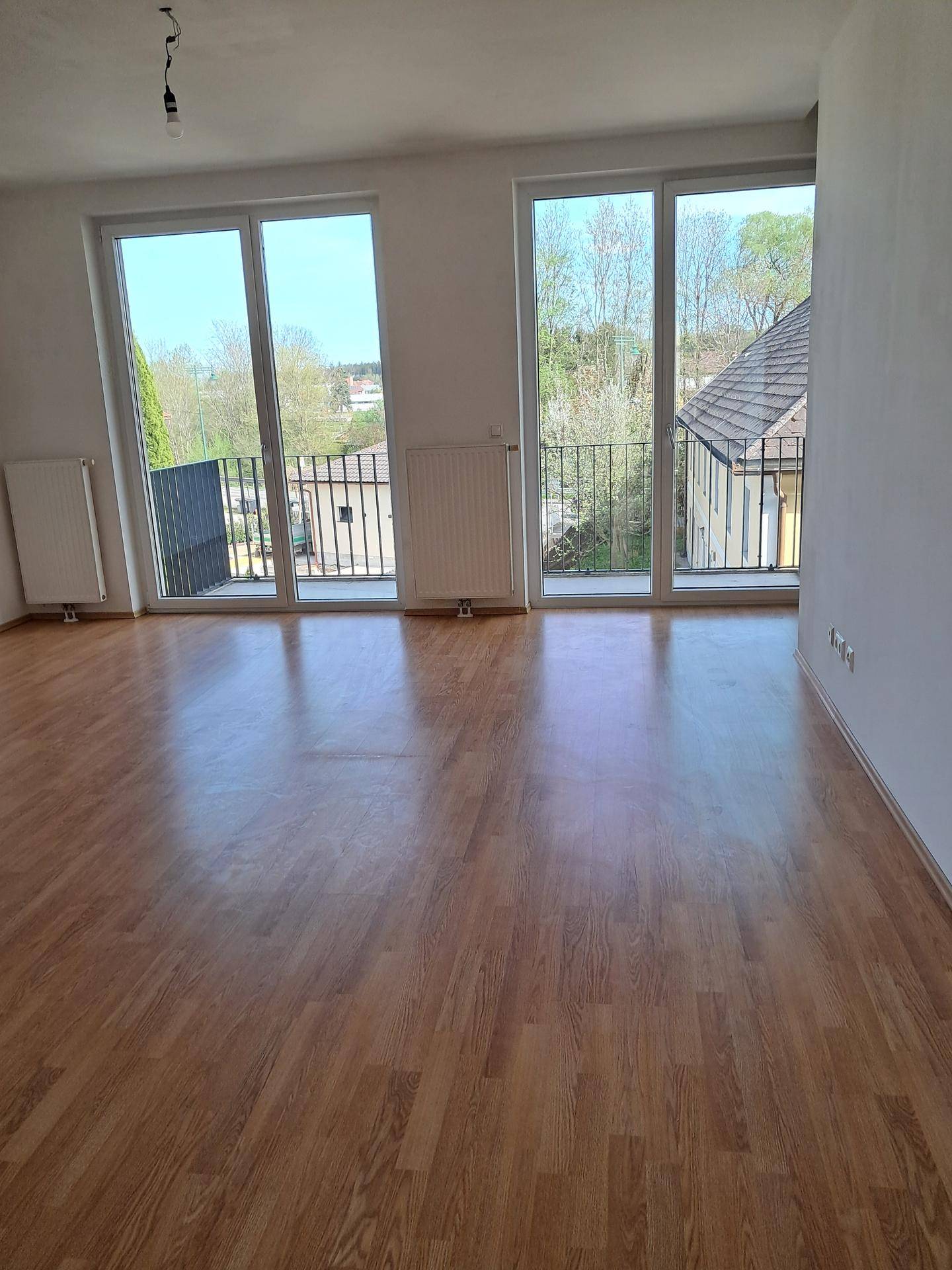 Heller Wohnbereich mit Laminatboden und großen Fenstern, die Zugang zu einem Balkon bieten.