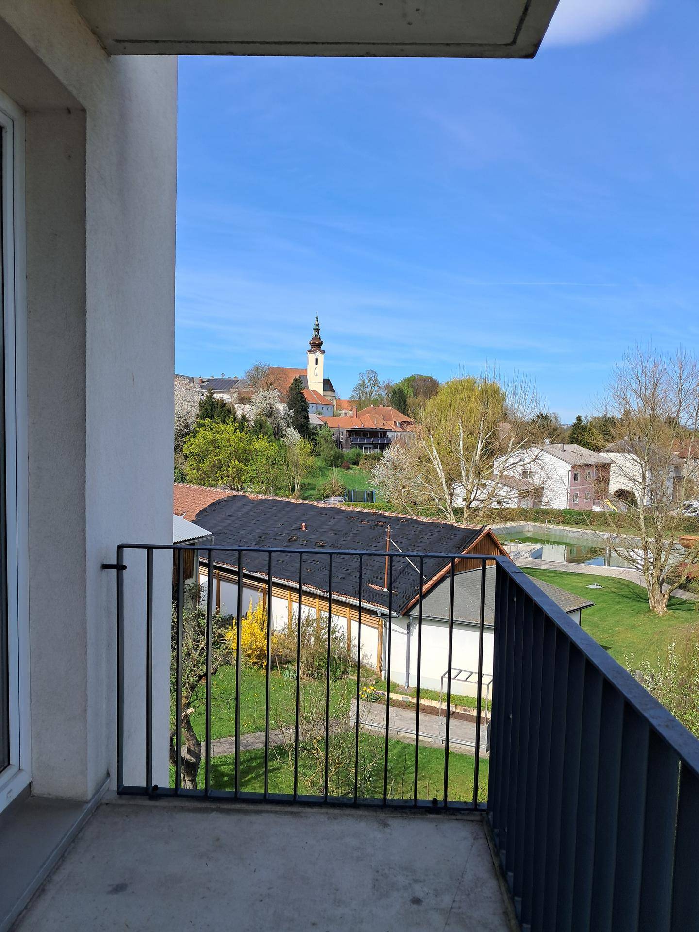 Balkon mit Blick auf eine grüne Umgebung und einen Kirchturm in der Ferne unter blauem Himmel.