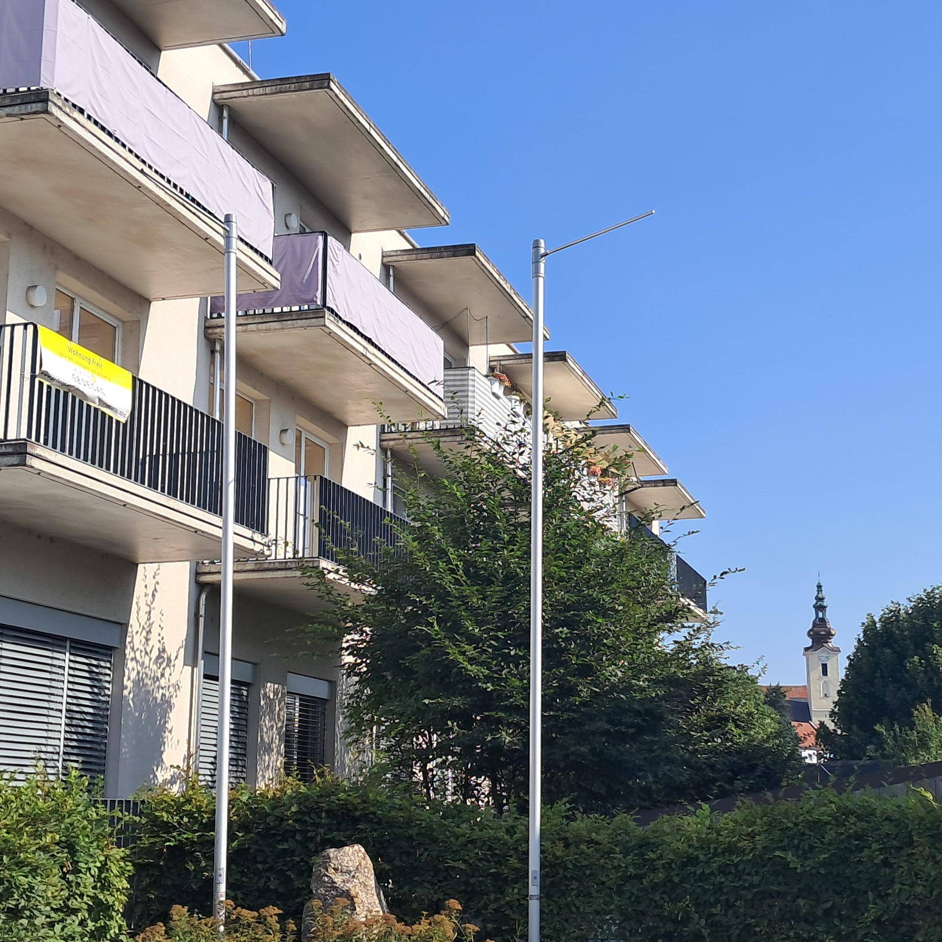 Fassade des modernen Gebäudes mit Balkonen und Rollläden, im Hintergrund ist ein Kirchturm sichtbar.