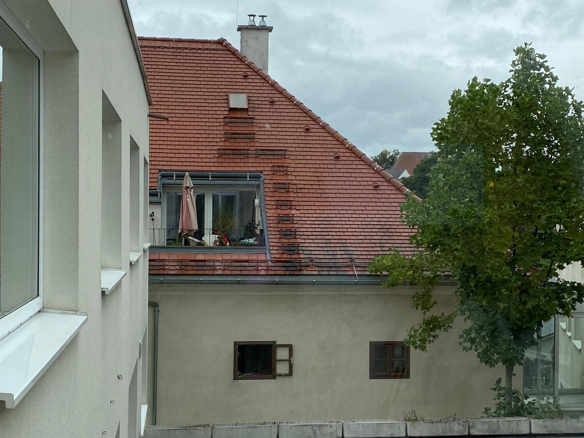 Blick aus dem Fenster auf ein rotes Ziegeldach und einen kleinen Balkon eines Nachbargebäudes.