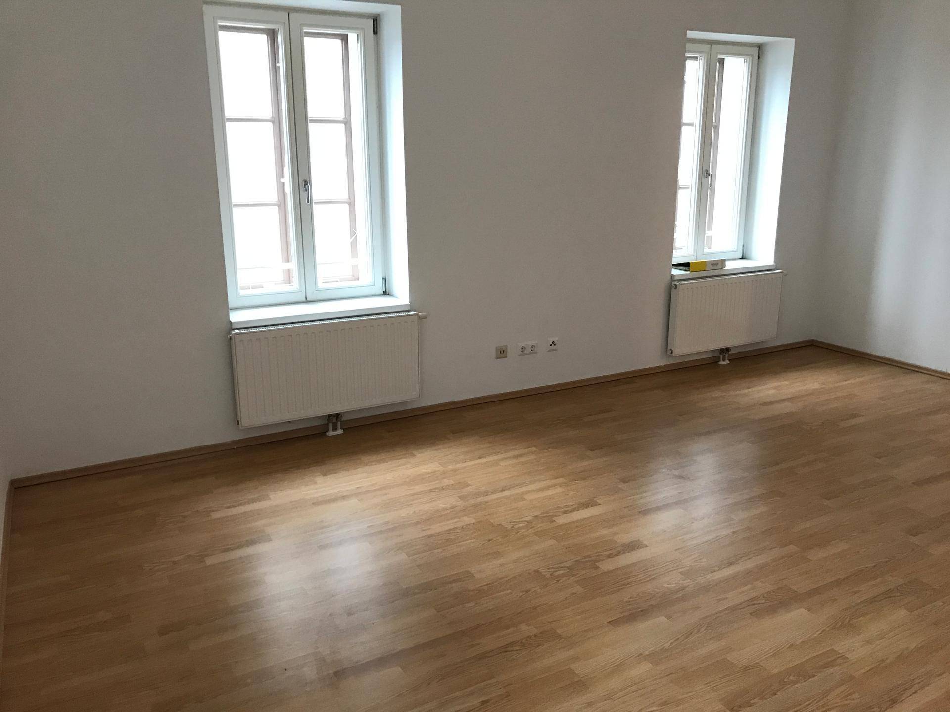 Geräumiges, leeres Wohnzimmer mit zwei Fenstern und glänzendem Laminatboden.