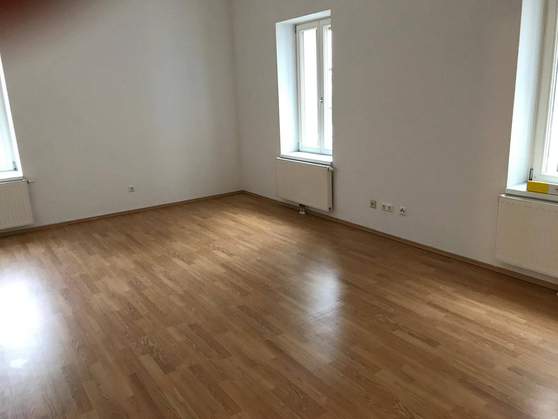 Helles, leeres Wohnzimmer mit zwei Fenstern und glänzendem Laminatboden.