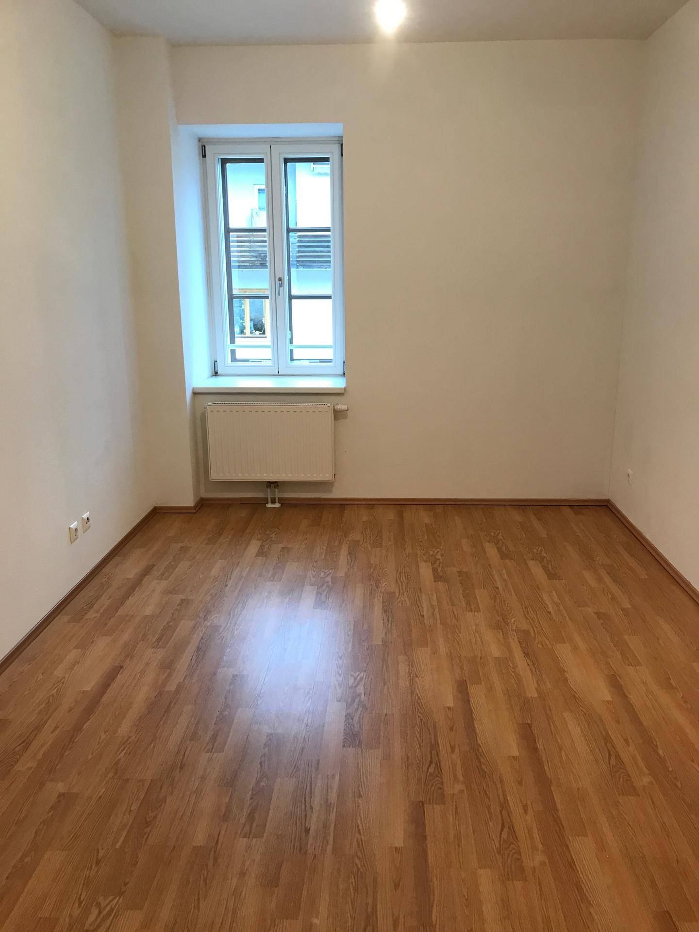 Helles, leeres Zimmer mit einem Fenster und glänzendem Laminatboden, bereit zur Einrichtung.