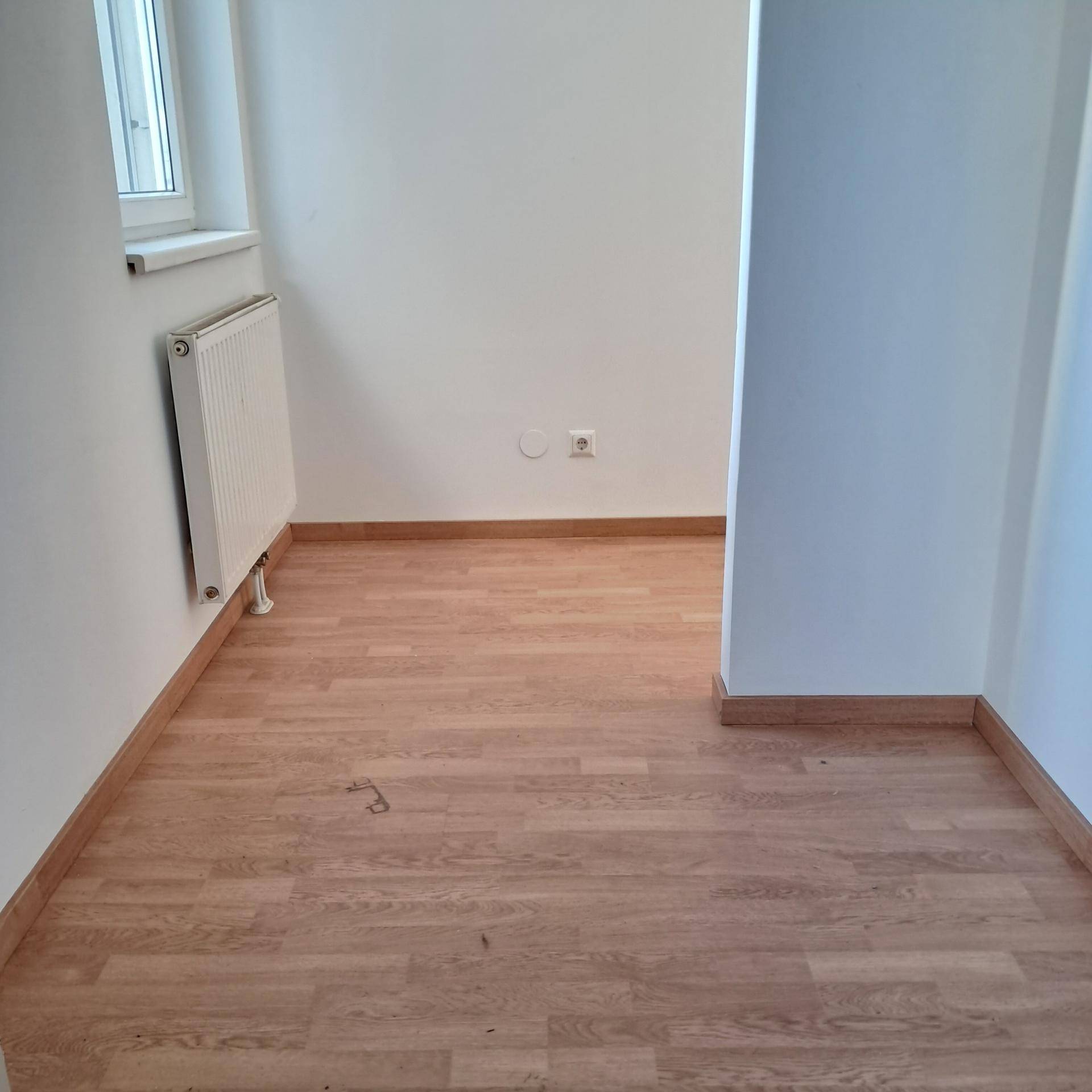 Leerer Raum mit hellem Laminatboden, weiß gestrichenen Wänden und einem Heizkörper unter dem Fenster.