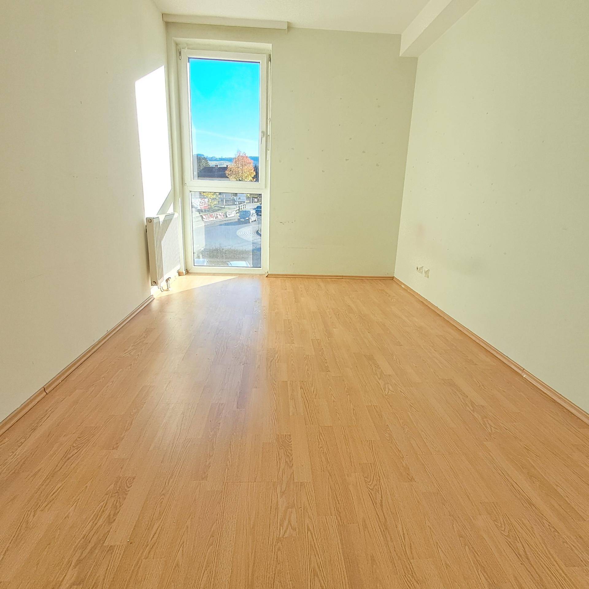 Leerer, heller Raum mit Laminatboden und einem großen Fenster, das viel Tageslicht hereinlässt.