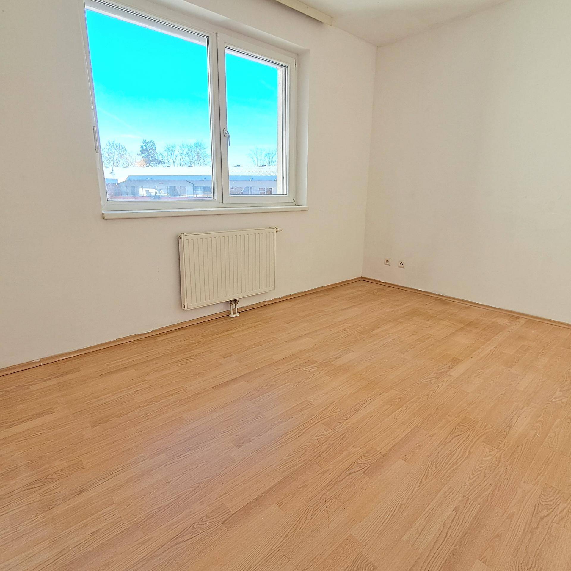 Heller Raum mit Laminatboden, weiß gestrichenen Wänden und einem Fenster mit Heizkörper darunter.