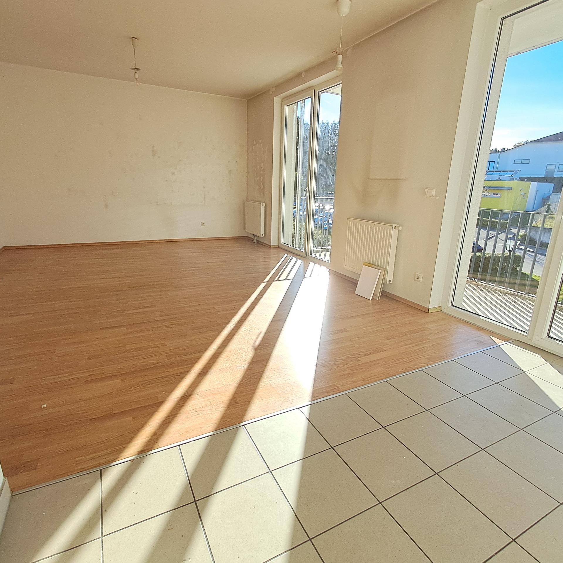 Heller Wohnbereich mit Laminatboden und Fliesen, großen Fenstern und Zugang zum Balkon.