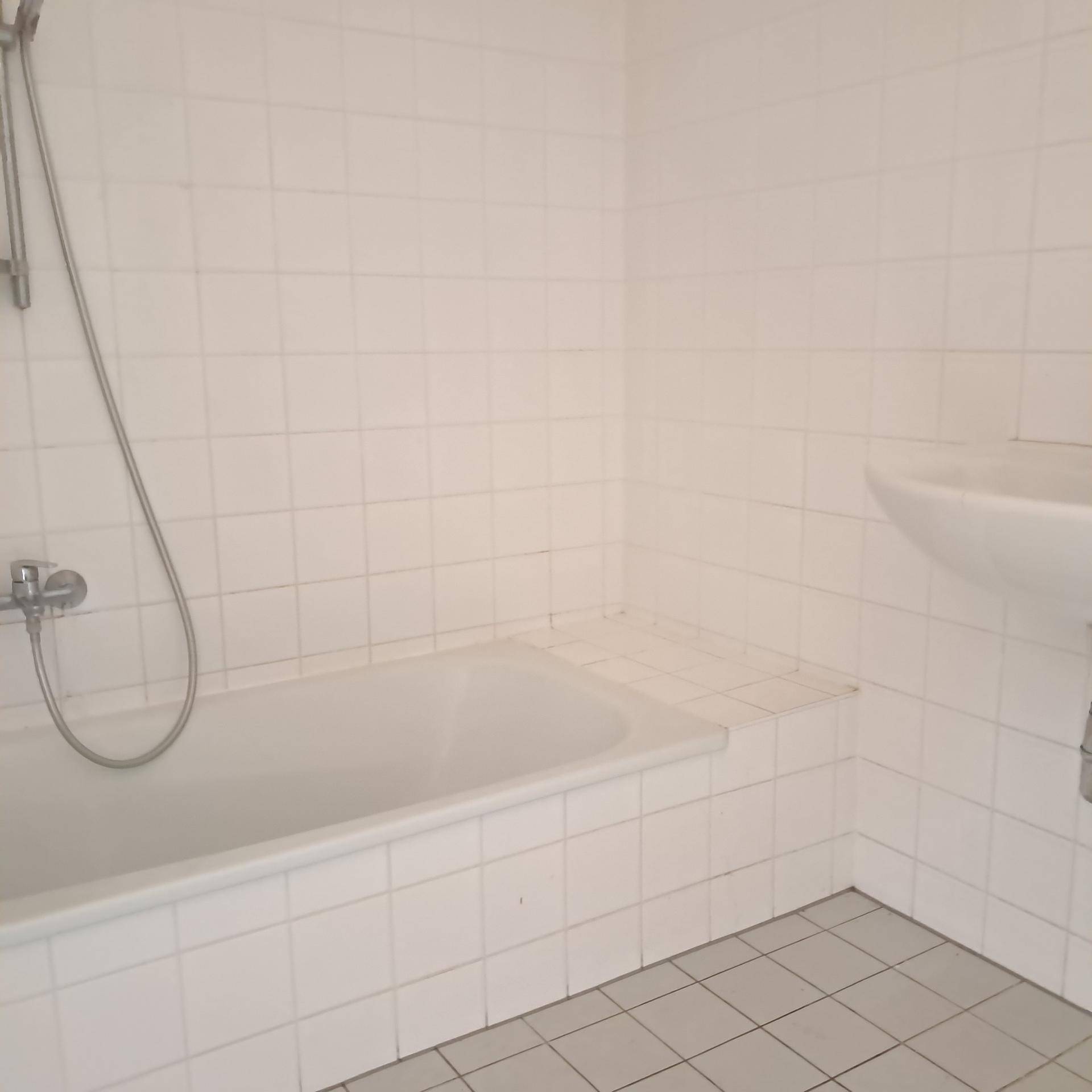 Badezimmer mit weißen Fliesen, einer Badewanne mit Duscharmatur und einem Waschbecken.