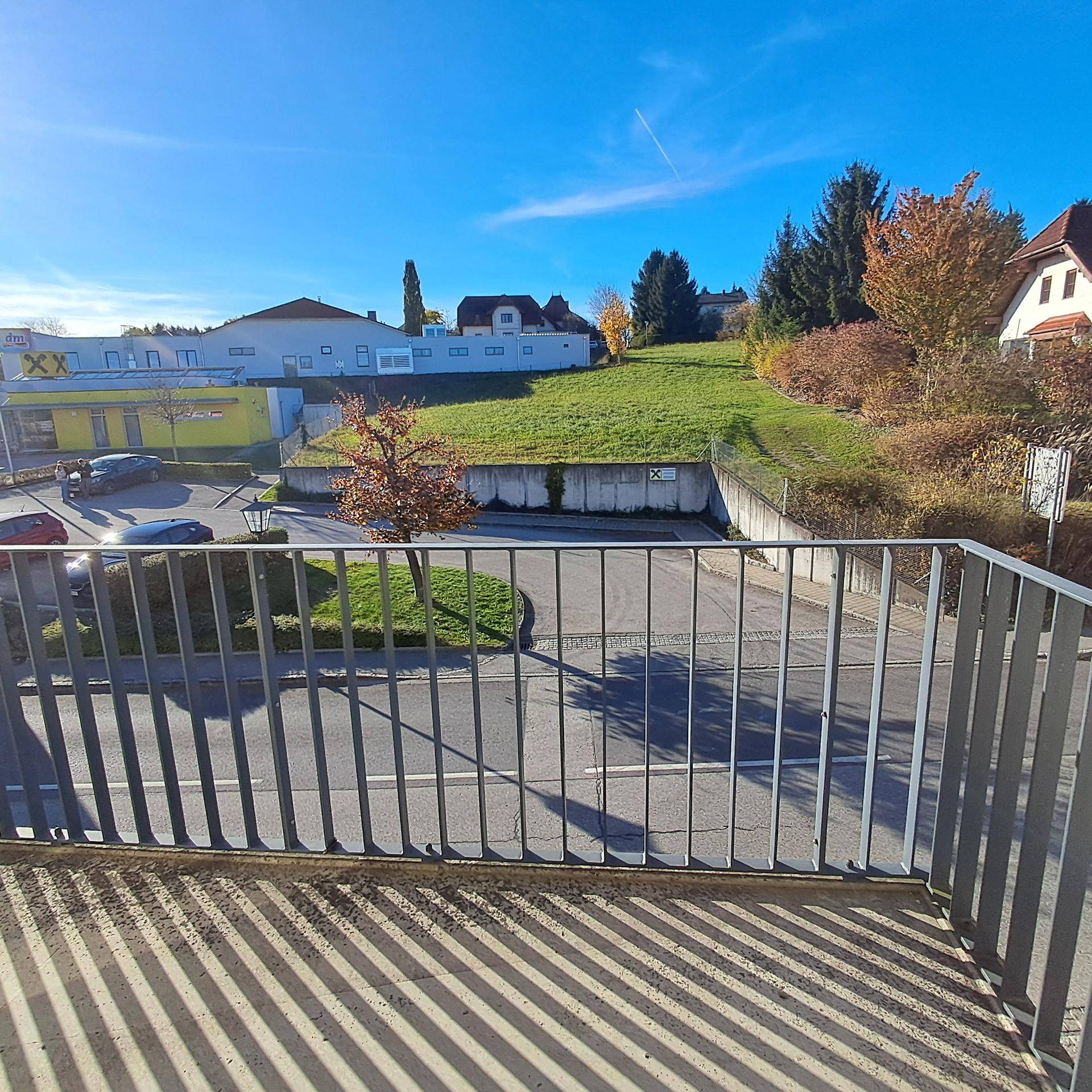 Blick vom Balkon auf die Straße, einen Parkplatz und eine grüne Hügellandschaft im Hintergrund.