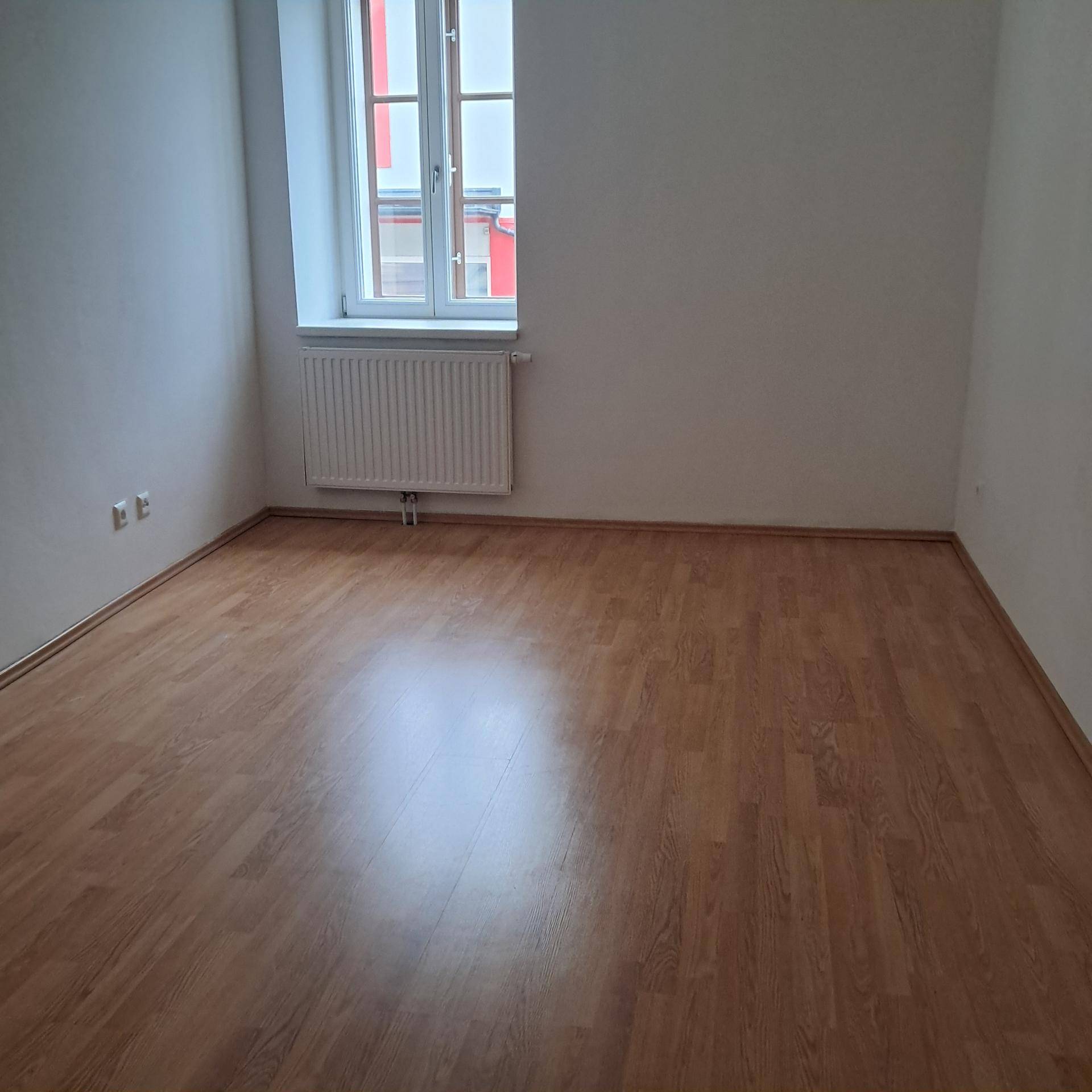 Leerer Raum mit hellem Laminatboden, weiß gestrichenen Wänden und einem Fenster.