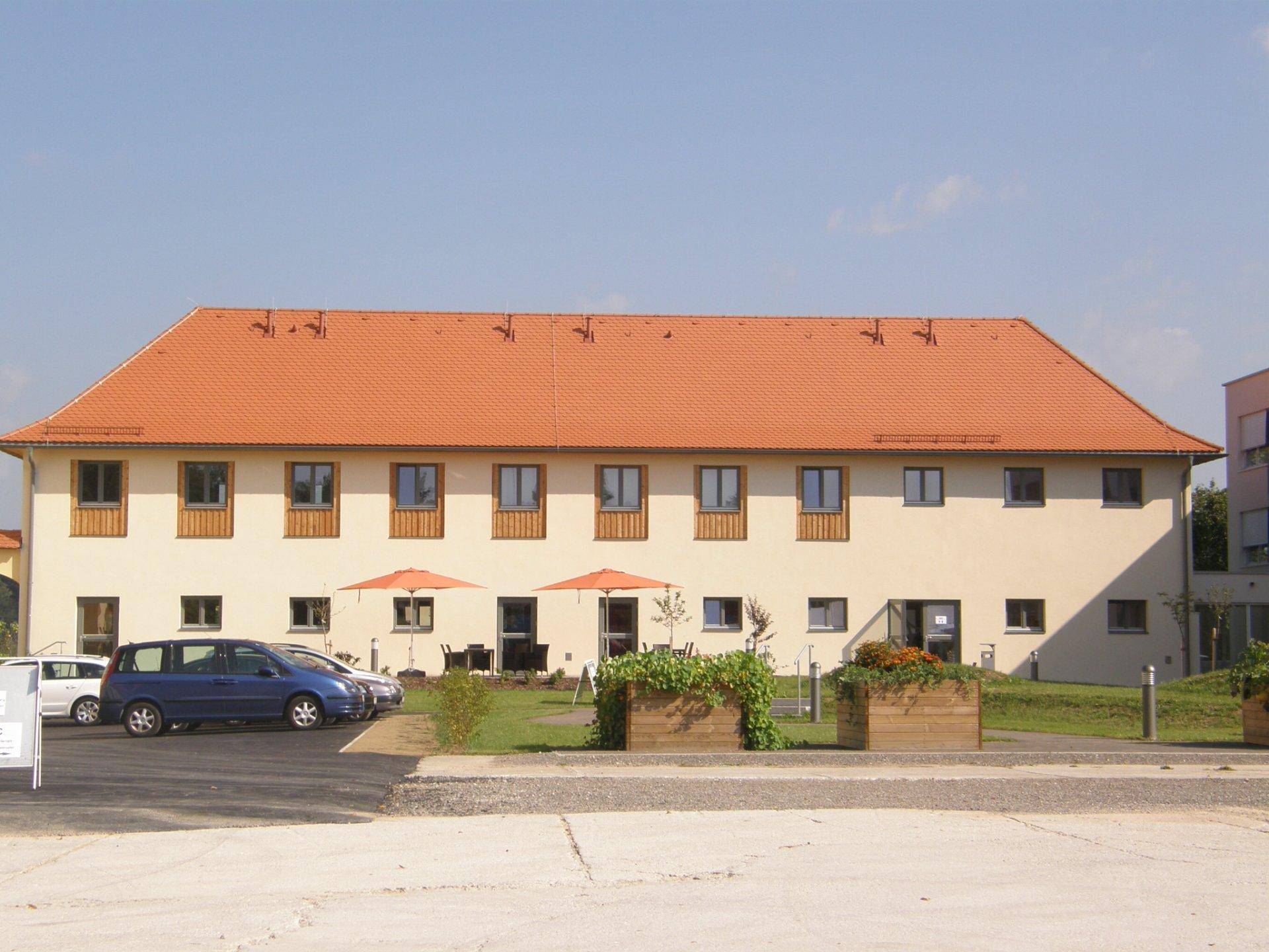 Längliches Wohngebäude mit rotem Ziegeldach, kleinen Balkonen und Außenbereich mit Sonnenschirmen.