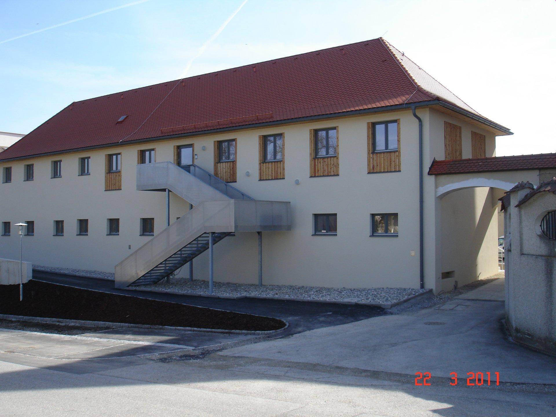 Seitenansicht eines Gebäudes mit rotem Ziegeldach und einer modernen Außentreppe aus Metall.