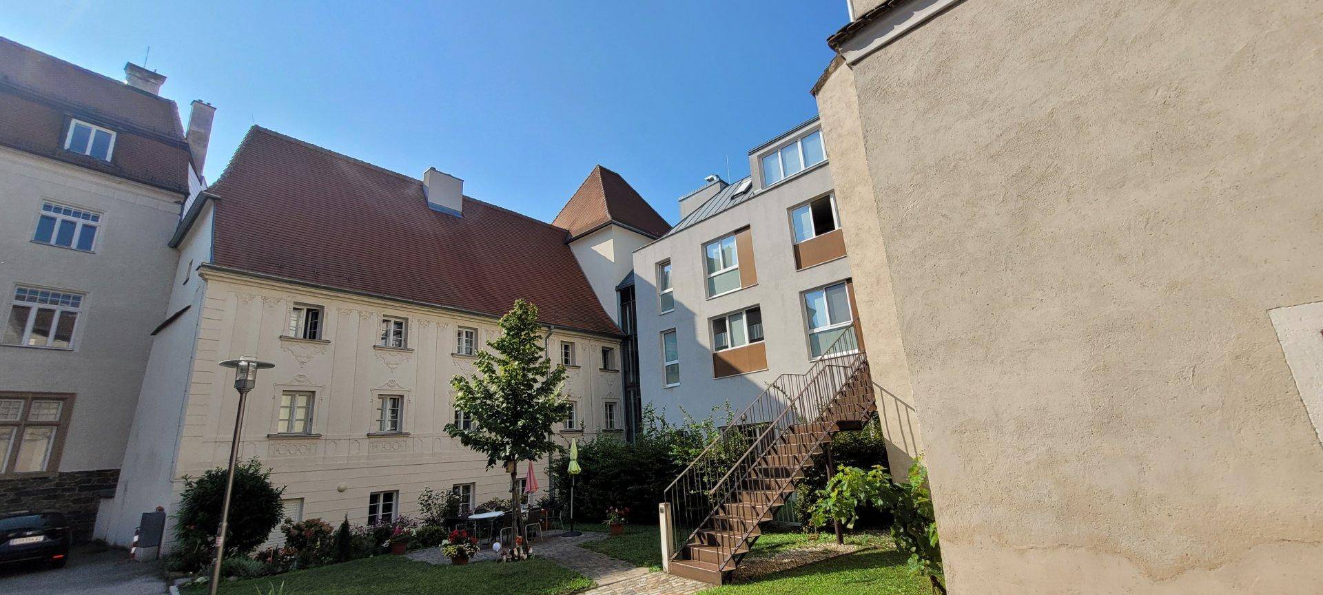 Sonniger Innenhof mit Grünflächen, Treppe und einer Mischung aus alten und neuen Gebäuden.
