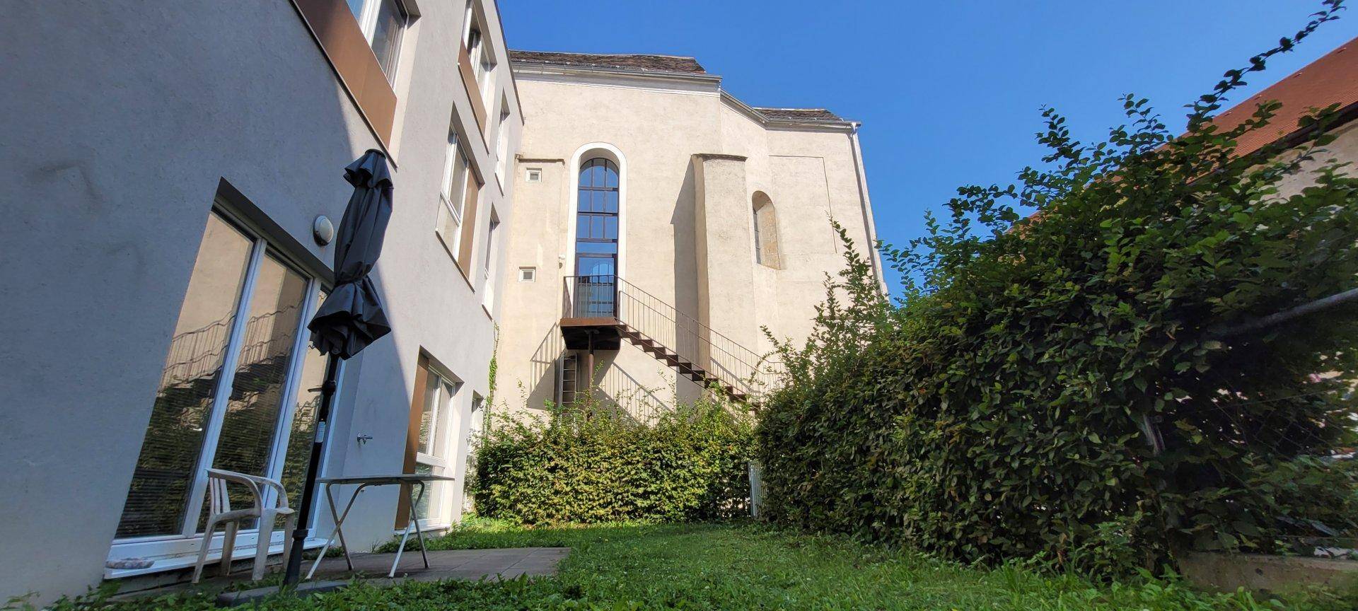 Kleiner Gartenbereich mit Hecke, Sitzgelegenheit und Blick auf ein historisches Gebäude.