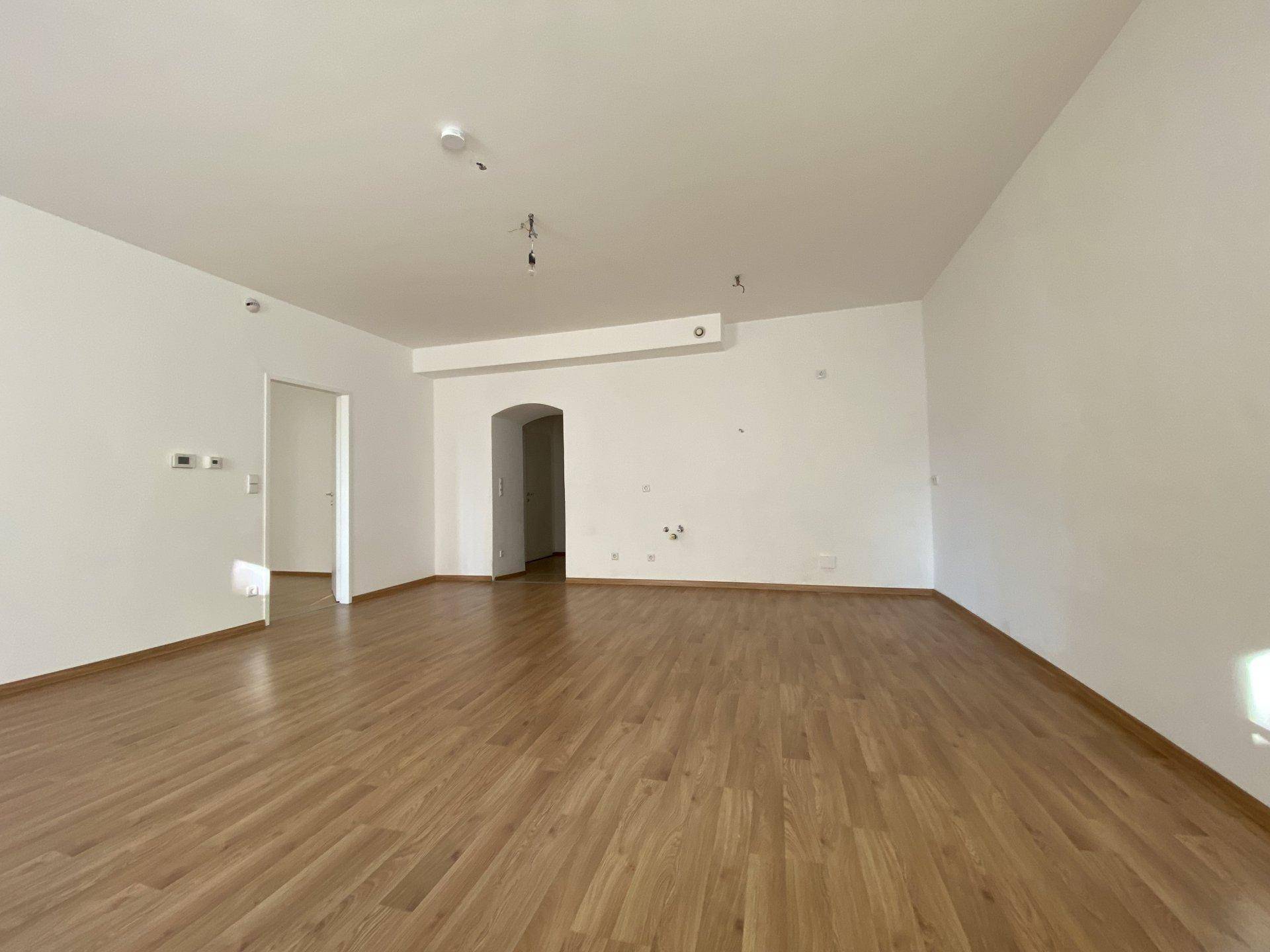 Geräumiges, leeres Wohnzimmer mit hellem Holzfußboden und weißen Wänden.