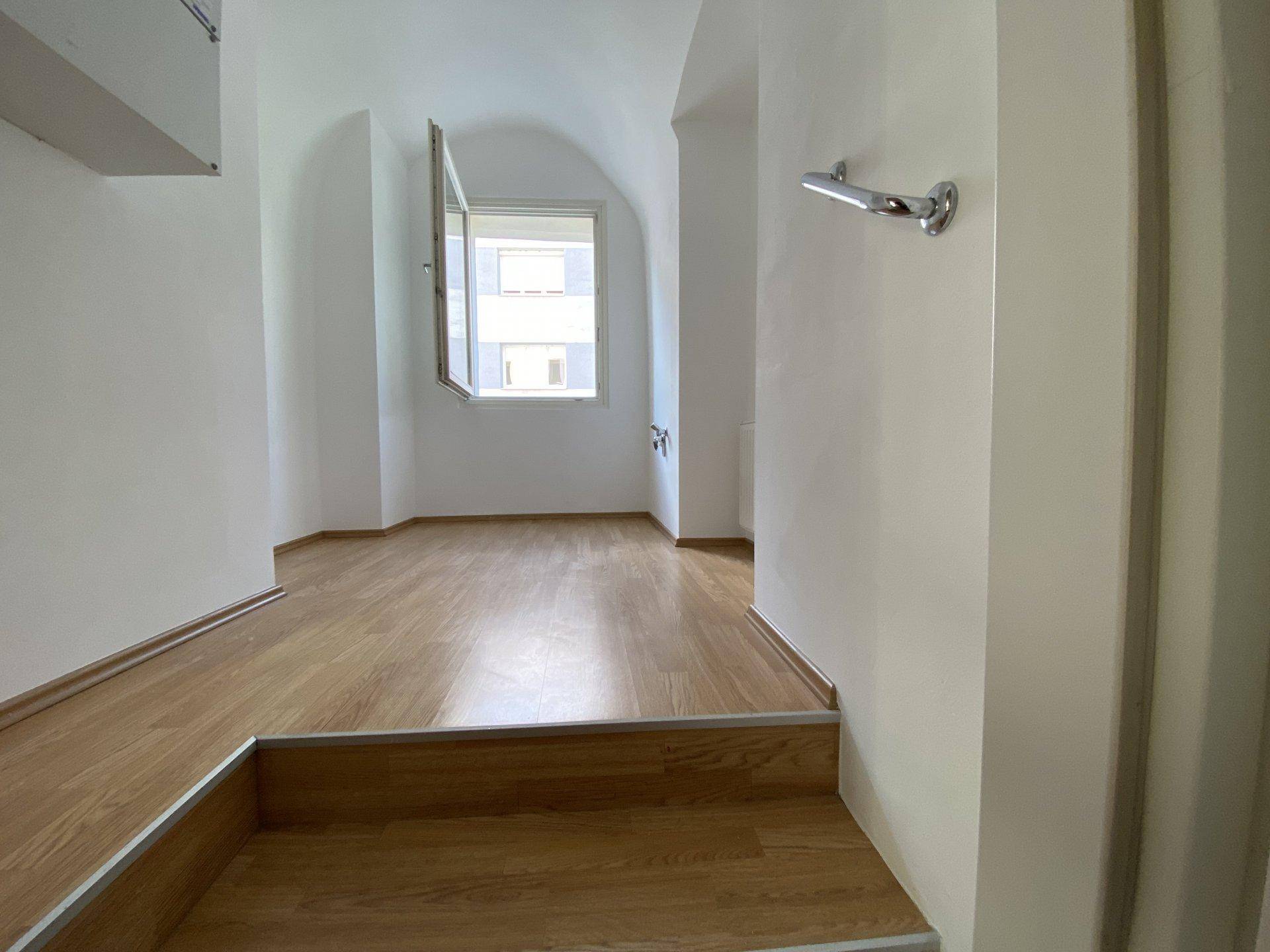 Kleiner Raum mit Holzfußboden, Fenster und einer Stufe, die den Bereich abgrenzt.