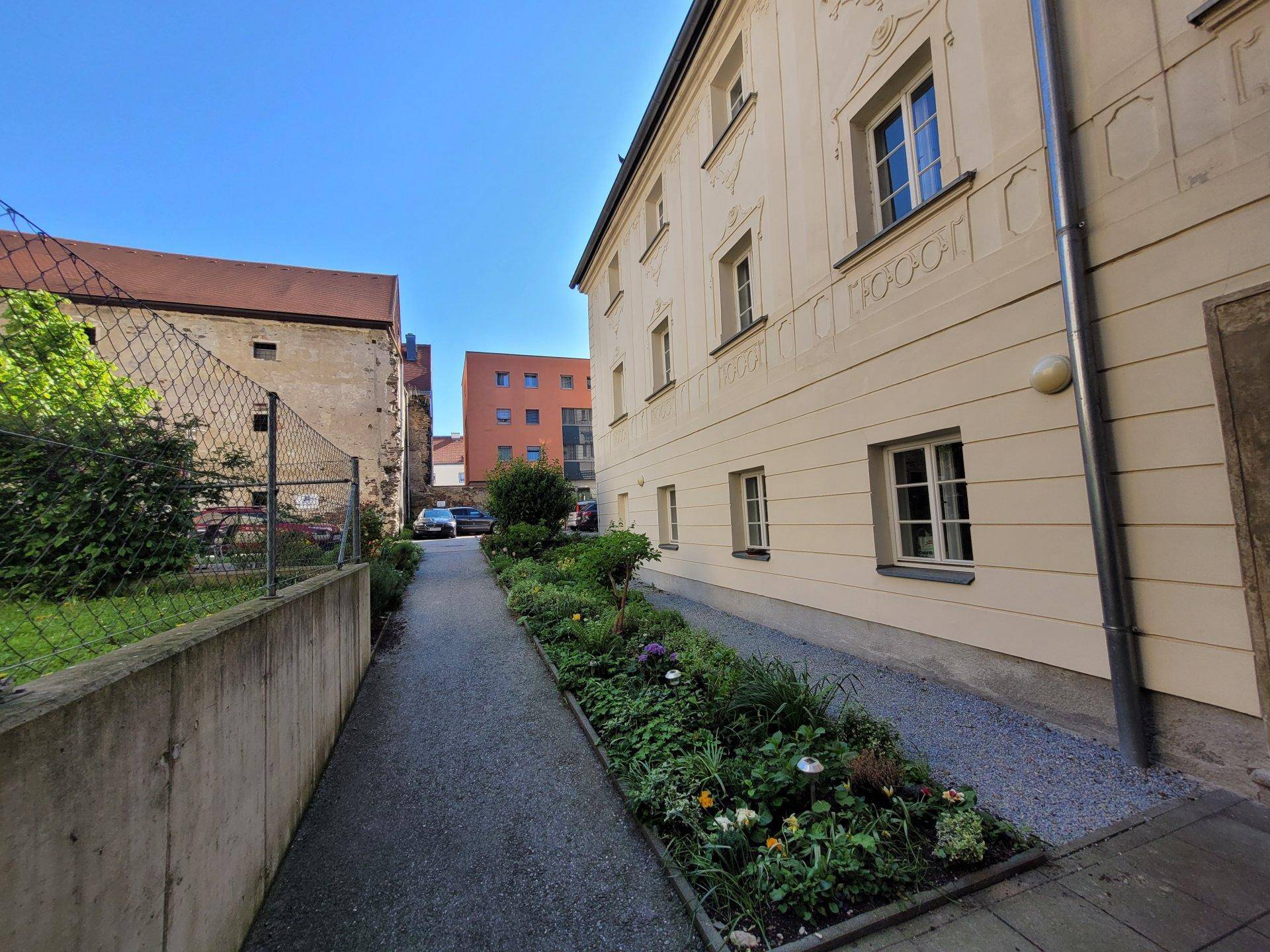 Schmaler Weg zwischen Gebäuden mit einem blühenden Blumenbeet und Kiesbelag.