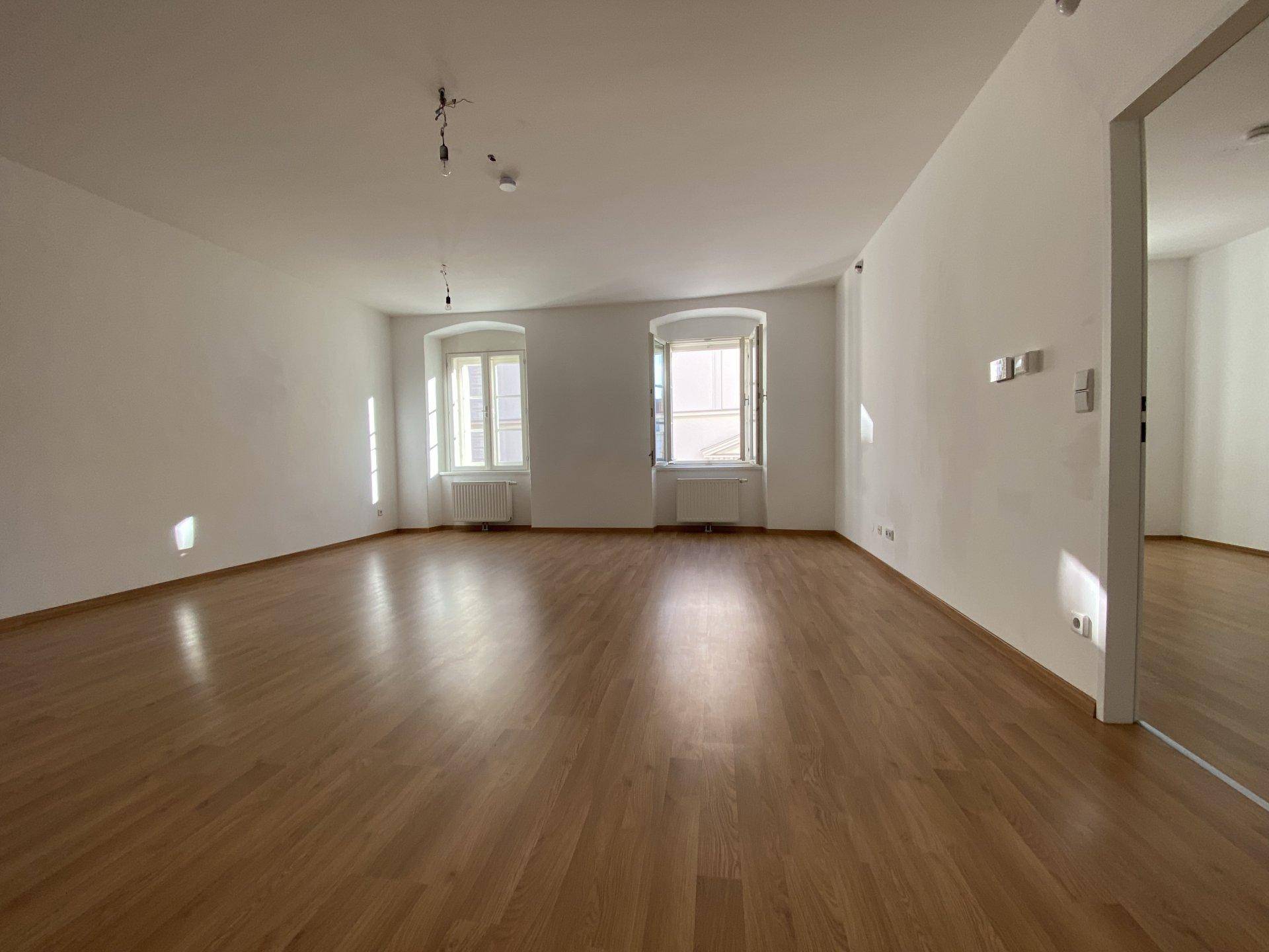 Helles und geräumiges Wohnzimmer mit zwei Fenstern und glänzendem Holzfußboden.